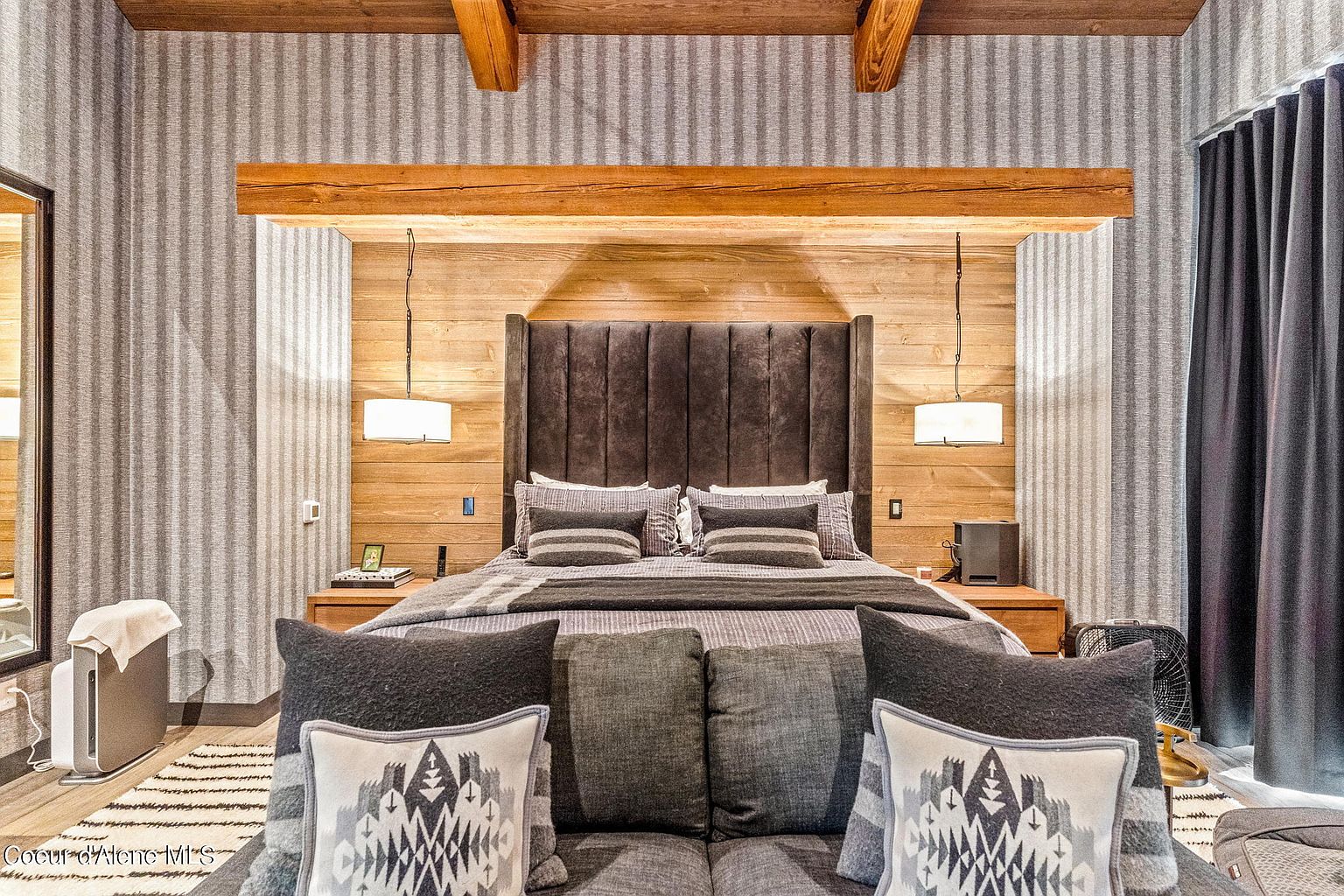This is a beautifully styled primary bedroom featuring a tall, upholstered headboard and a neutral color palette. The room has wood paneling behind the bed, complemented by wooden beams on the ceiling. The bedding includes multiple pillows and a textured throw, creating a cozy and inviting atmosphere.