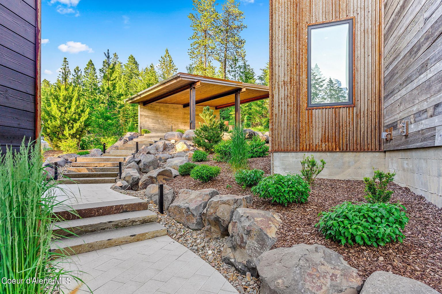 This image showcases a beautifully landscaped yard with a modern architectural style. Stone steps lead up to a covered outdoor structure, surrounded by lush greenery and carefully placed rocks. The exterior of the building features a mix of materials, including wood and corrugated metal, creating a unique and appealing aesthetic.