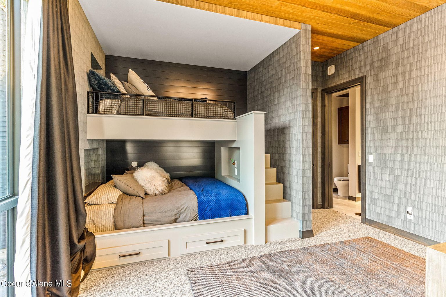 This is an interior shot of a guest bedroom featuring a custom-built bunk bed with integrated storage drawers and stairs. The room is decorated with a neutral color palette, including gray and white tones, and the walls are covered in a textured wallpaper. The overall impression is modern and cozy, making it an inviting space for guests.