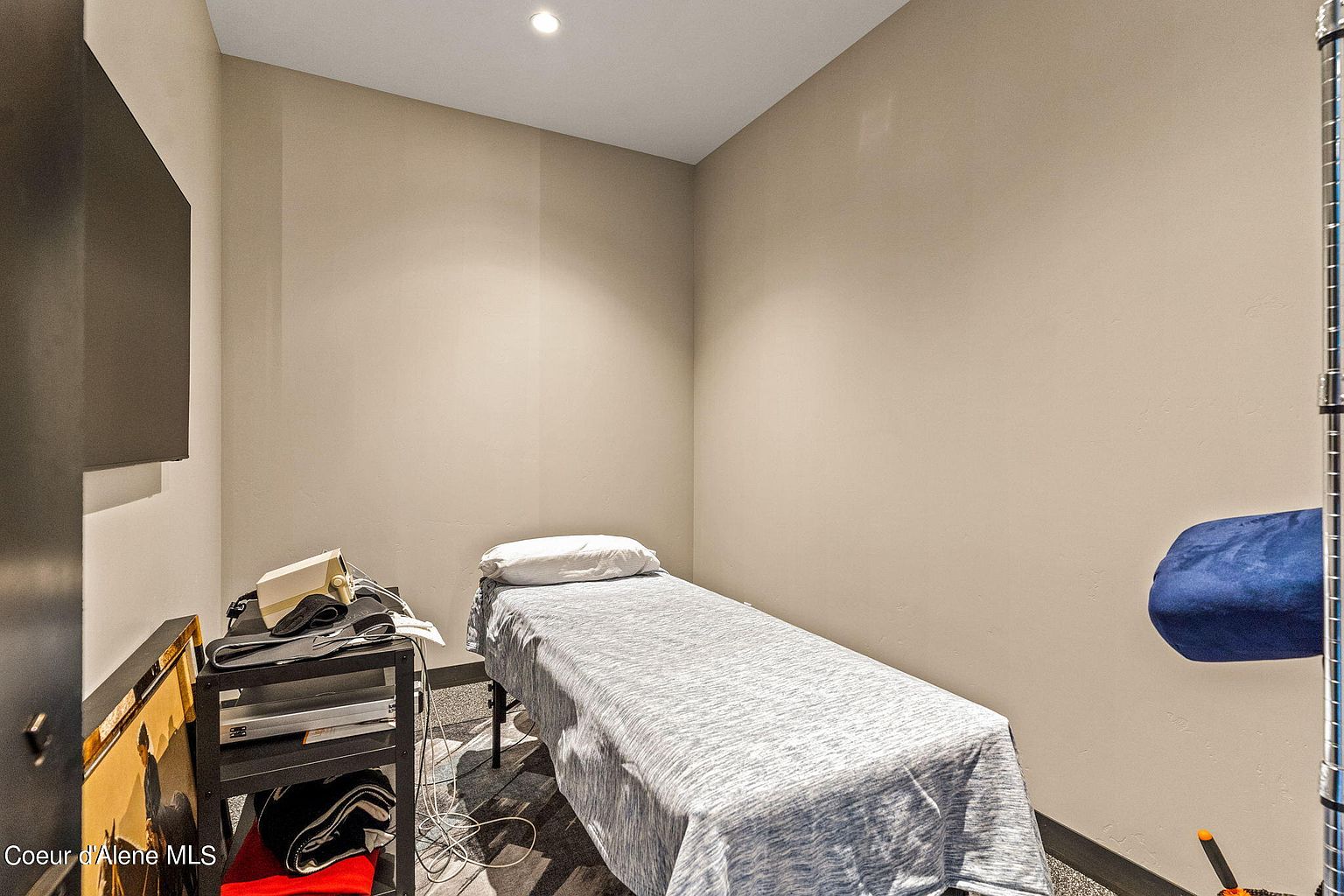 This is a simple, functional bedroom featuring a single bed with a light blue and white patterned coverlet and a white pillow. A small black shelving unit holds various items, including a white device and some folded clothes. The walls are painted in a neutral beige tone, and the flooring appears to be dark gray carpet, creating a minimalist and clean aesthetic.