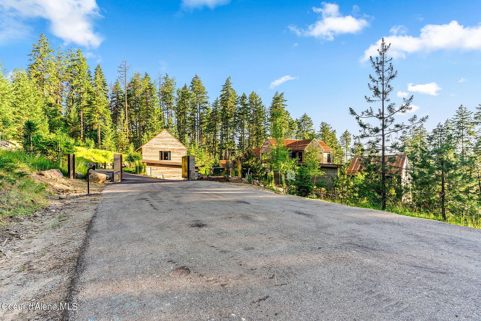 This image showcases the front view of a rustic property nestled among tall trees. The property features wooden structures with a mix of new and aged elements, including a gatehouse and a main house with a visible roof. The driveway leads up to the buildings, creating a sense of seclusion and natural beauty.