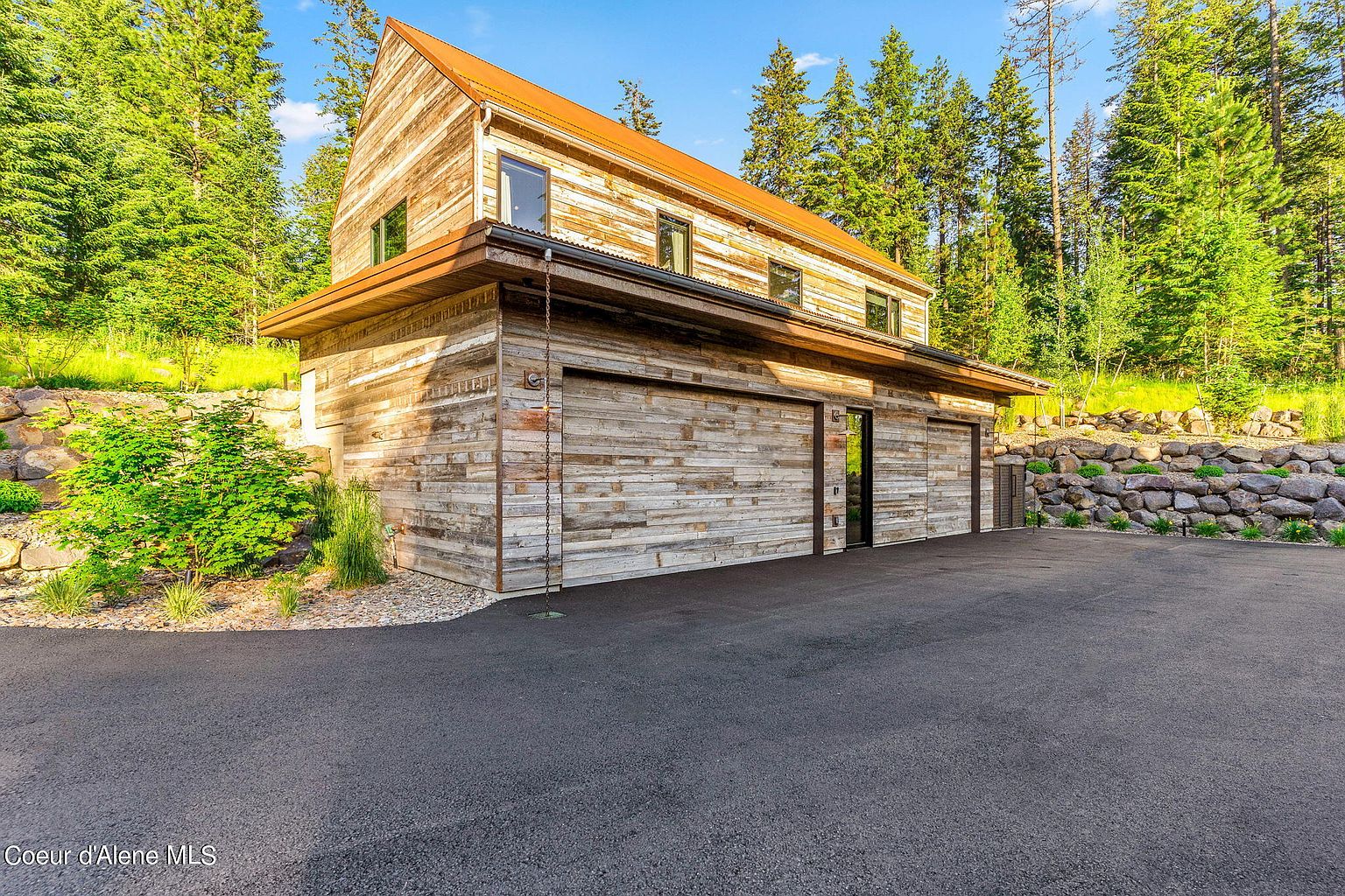This image showcases the exterior of a rustic-style garage, featuring wooden plank siding and a metal roof. The garage has multiple bays and a dark asphalt driveway. The surrounding landscape includes mature trees and a stone retaining wall, adding to the property's natural appeal.