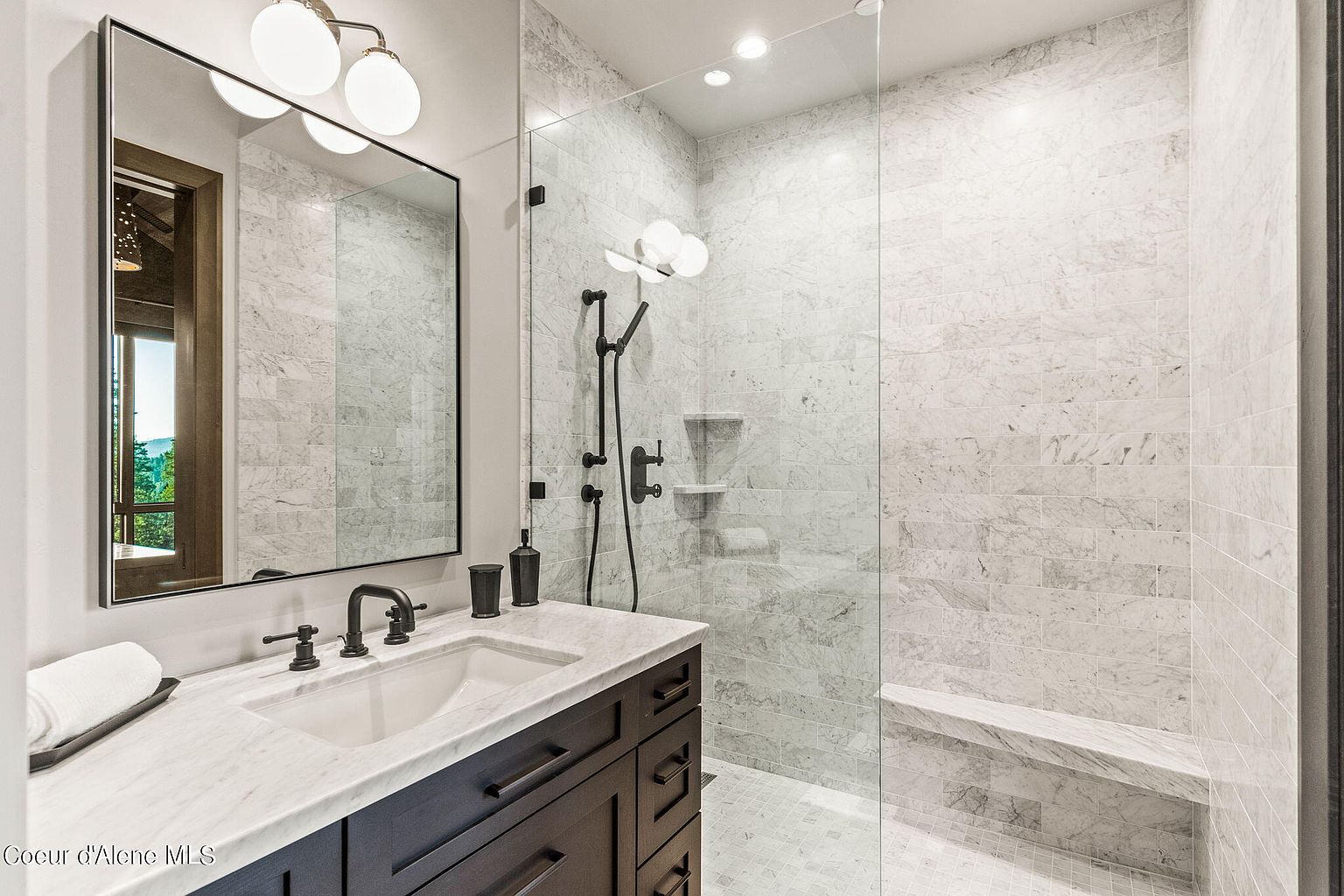 This is a primary bathroom featuring a modern design with marble tiling throughout the shower and vanity area. The vanity has a dark wood cabinet with a white marble countertop and a rectangular sink. A large mirror hangs above the vanity, reflecting natural light, and the shower is enclosed with glass, showcasing a built-in bench and sleek black fixtures. The overall impression is luxurious and spa-like.
