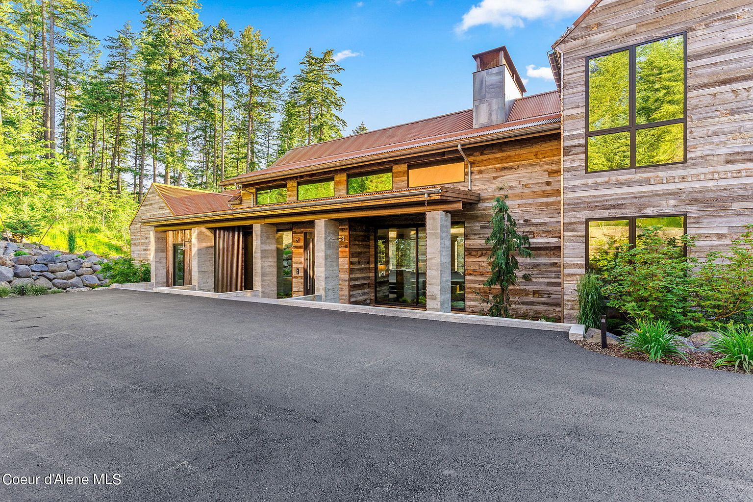 This is a front exterior view of a modern rustic home. The house features a combination of wood siding and stone accents, with large windows reflecting the surrounding trees. A wide driveway leads up to the entrance, and the landscaping includes mature trees and shrubs, creating a sense of privacy and natural beauty.