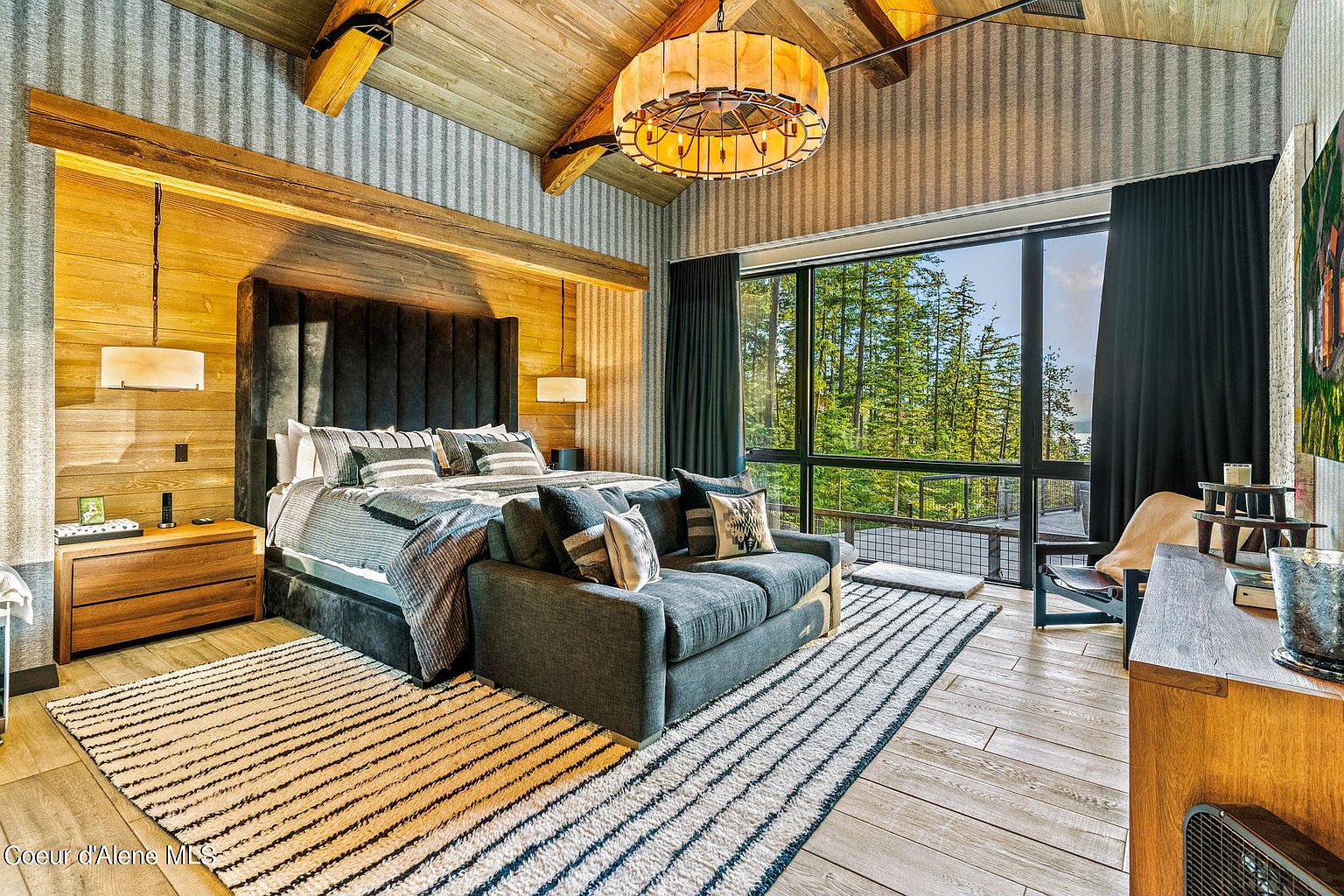 This is a luxurious primary bedroom featuring a king-size bed with a dark upholstered headboard and high-end bedding. A small sofa sits at the foot of the bed, and a large window offers a view of the surrounding forest. The room has a rustic-modern style with wood paneling and a unique chandelier.
