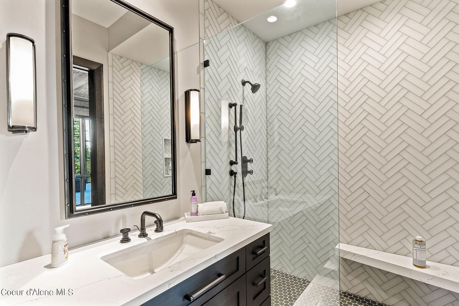 This is a primary bathroom featuring a modern design with a dark wood vanity topped with a white countertop and a rectangular sink. A large mirror hangs above the vanity, flanked by sconce lighting. The walk-in shower is enclosed with glass and features white herringbone tile and a built-in bench, creating a luxurious and spa-like atmosphere.