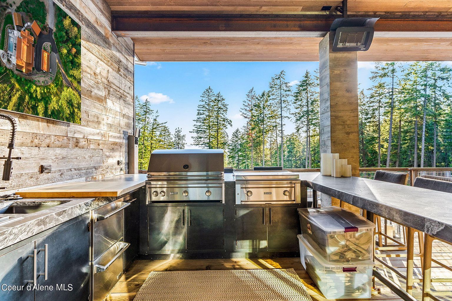 This image showcases an outdoor kitchen area on a patio or deck, featuring stainless steel appliances, including a grill and a side burner, built into dark cabinetry. A wooden countertop extends to a bar area with seating, and clear storage bins are neatly stacked underneath. The backdrop reveals a scenic view of tall evergreen trees under a bright blue sky, creating a luxurious and inviting outdoor living space.