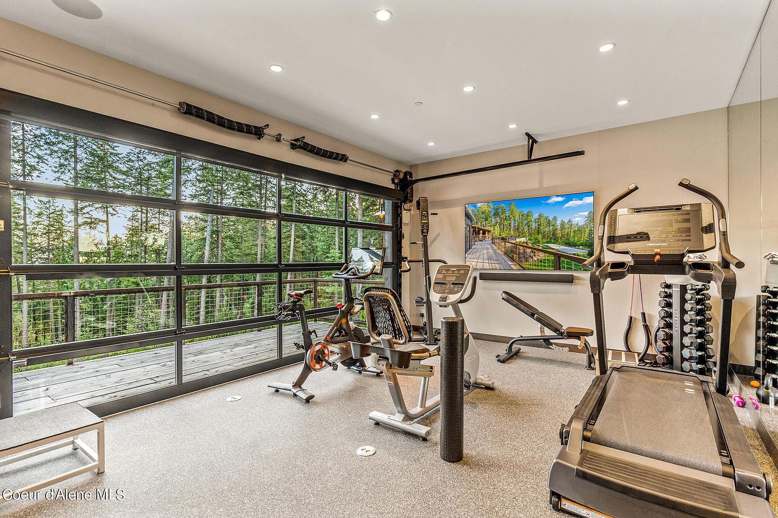 This is a well-equipped home gym featuring a treadmill, stationary bikes, and weight training equipment. A large glass door provides ample natural light and a view of the surrounding trees, creating an inviting workout environment. The room has a neutral color palette and a rubberized floor, enhancing both safety and aesthetics.