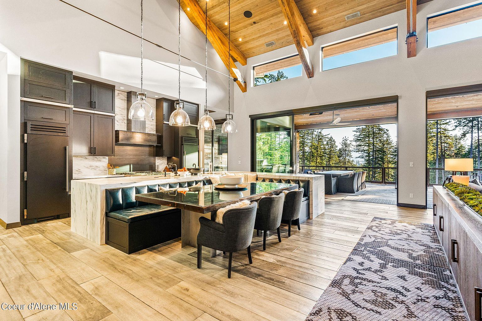 This is a spacious, modern kitchen with dark cabinetry, marble countertops, and a large island that doubles as a dining area with booth seating. The kitchen features pendant lighting, stainless steel appliances, and an open floor plan that leads to an outdoor deck with views of the surrounding trees. The high ceilings with exposed wooden beams add a touch of rustic charm to the contemporary design.