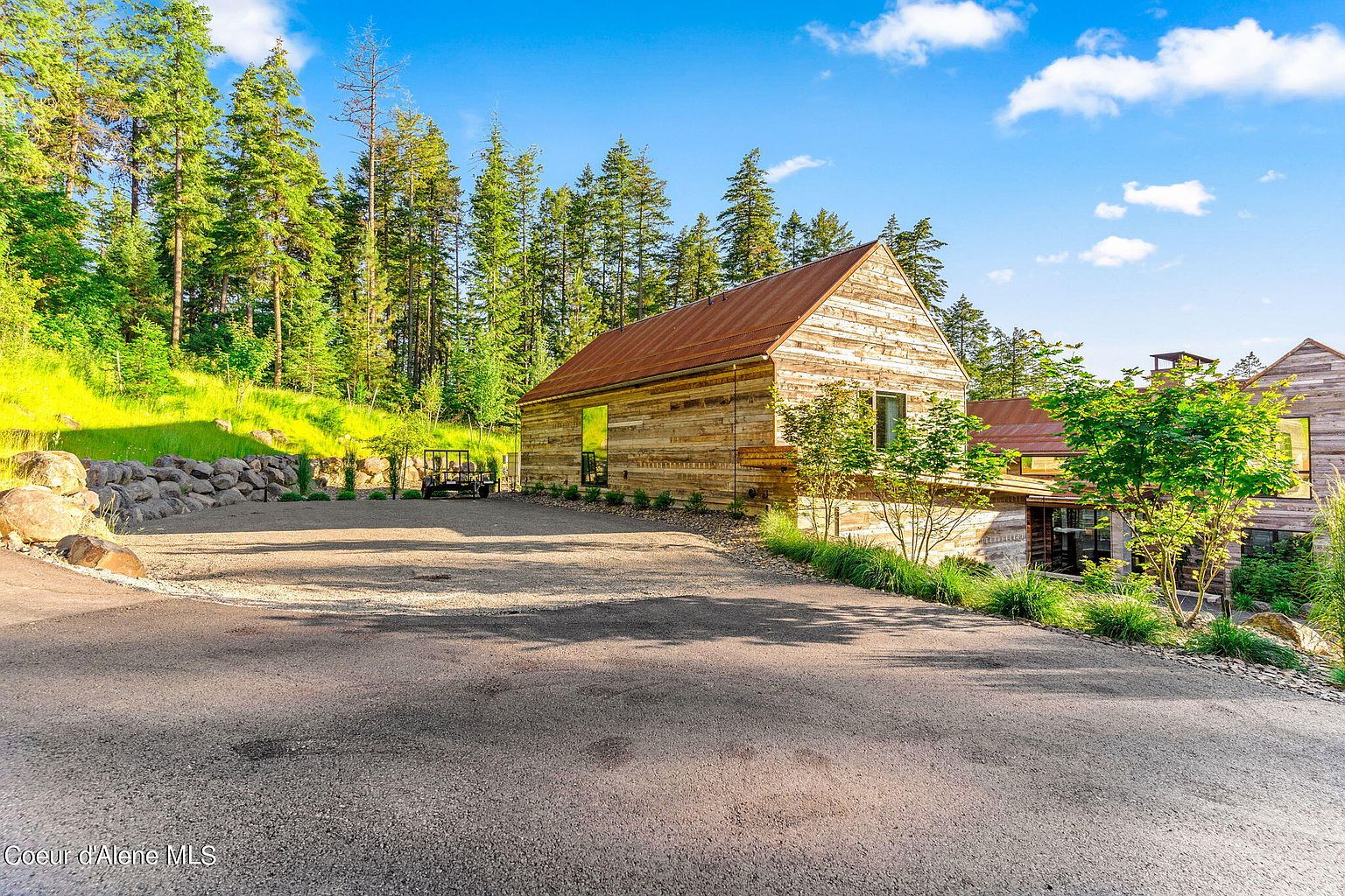 This image showcases the front exterior of a rustic-style home with wooden siding and a metal roof. The house is nestled among tall trees and greenery, creating a serene and private setting. A spacious driveway leads up to the house, offering ample parking space.