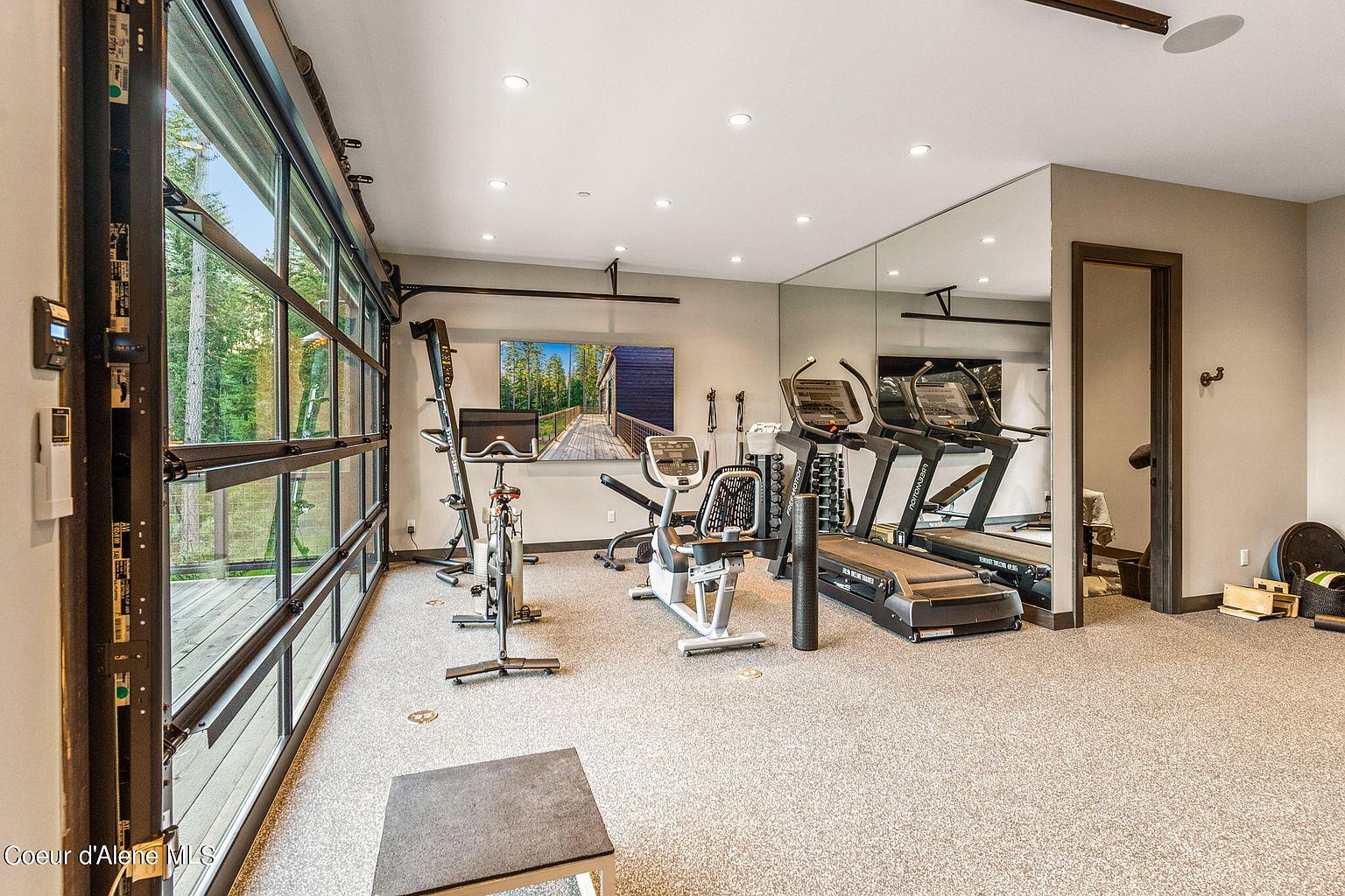 This is an interior shot of a well-equipped home gym. The room features a variety of exercise equipment, including treadmills, stationary bikes, and a weight rack. A large mirror covers one wall, enhancing the sense of space, while a glass garage door provides natural light and a view of the outdoors. The perspective is from a medium distance, showcasing the layout and equipment.