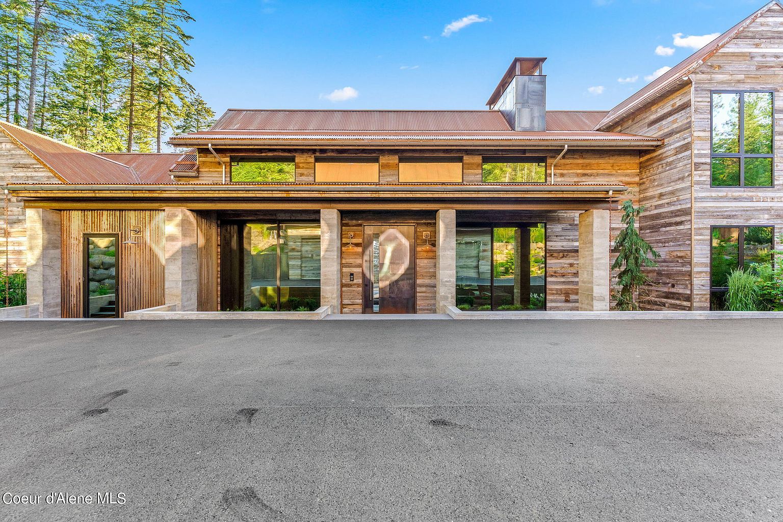 This is a front view of a modern rustic home. The exterior features a combination of wood and stone, with large windows and a metal roof. The entryway is framed by stone columns, and the driveway leads up to the house. The overall impression is one of luxury and sophistication.
