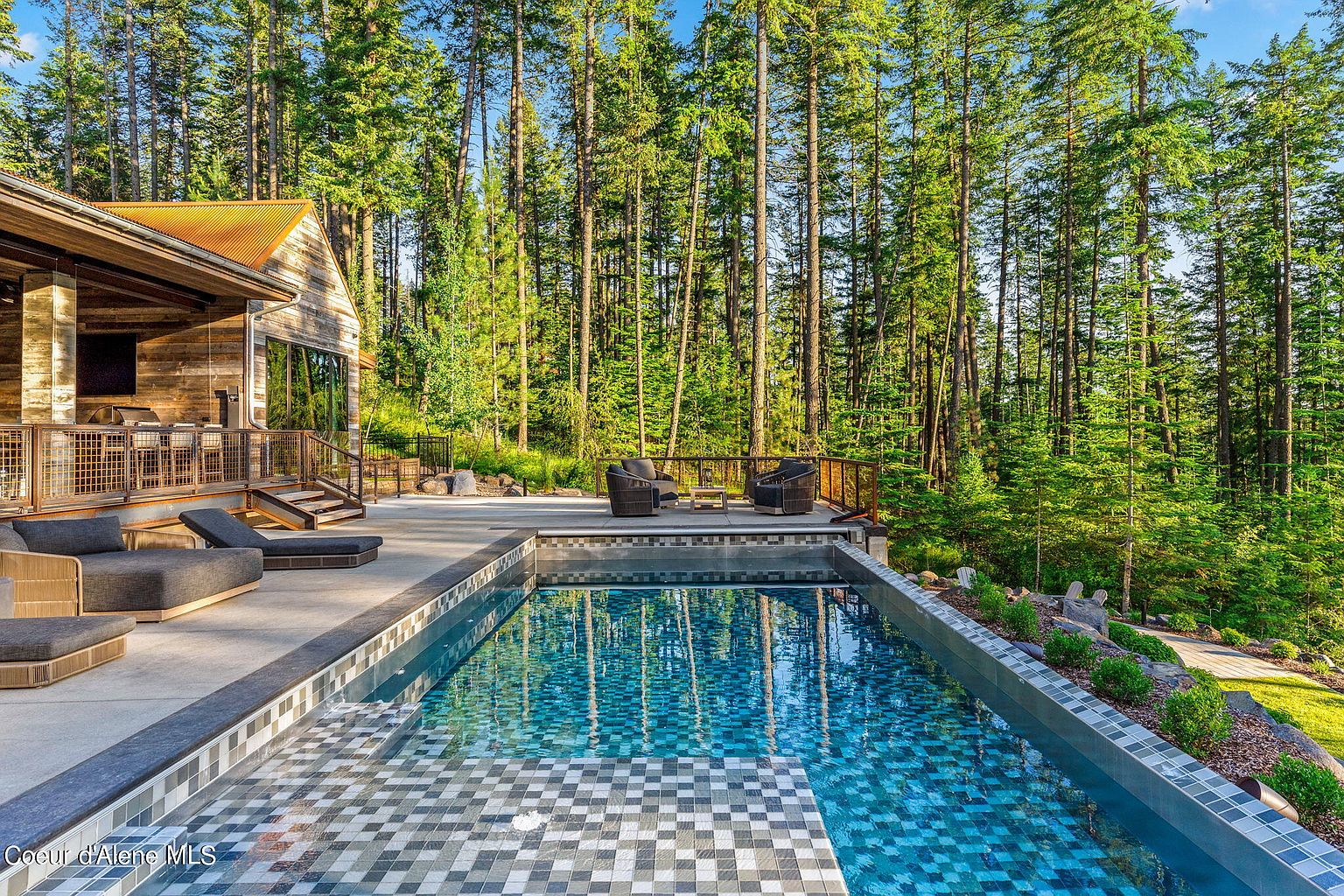 This image showcases a luxurious outdoor pool area, featuring a modern pool with intricate tile work and clear blue water. The pool is surrounded by a spacious patio with comfortable lounge furniture and a seating area with stylish chairs. Tall trees provide a scenic backdrop, creating a serene and private atmosphere.
