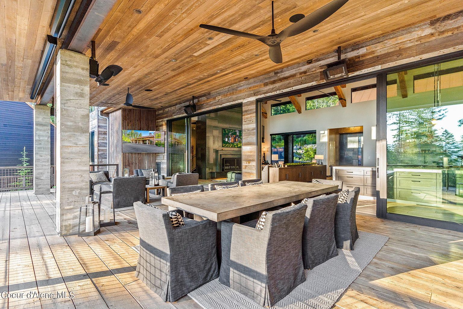 This image showcases a luxurious outdoor patio area, featuring a large wooden dining table surrounded by upholstered chairs, perfect for entertaining. The space is covered by a wooden ceiling with ceiling fans and includes a comfortable lounge area with a TV, creating a seamless transition between indoor and outdoor living. Large sliding glass doors offer views of the surrounding landscape and provide access to the interior.