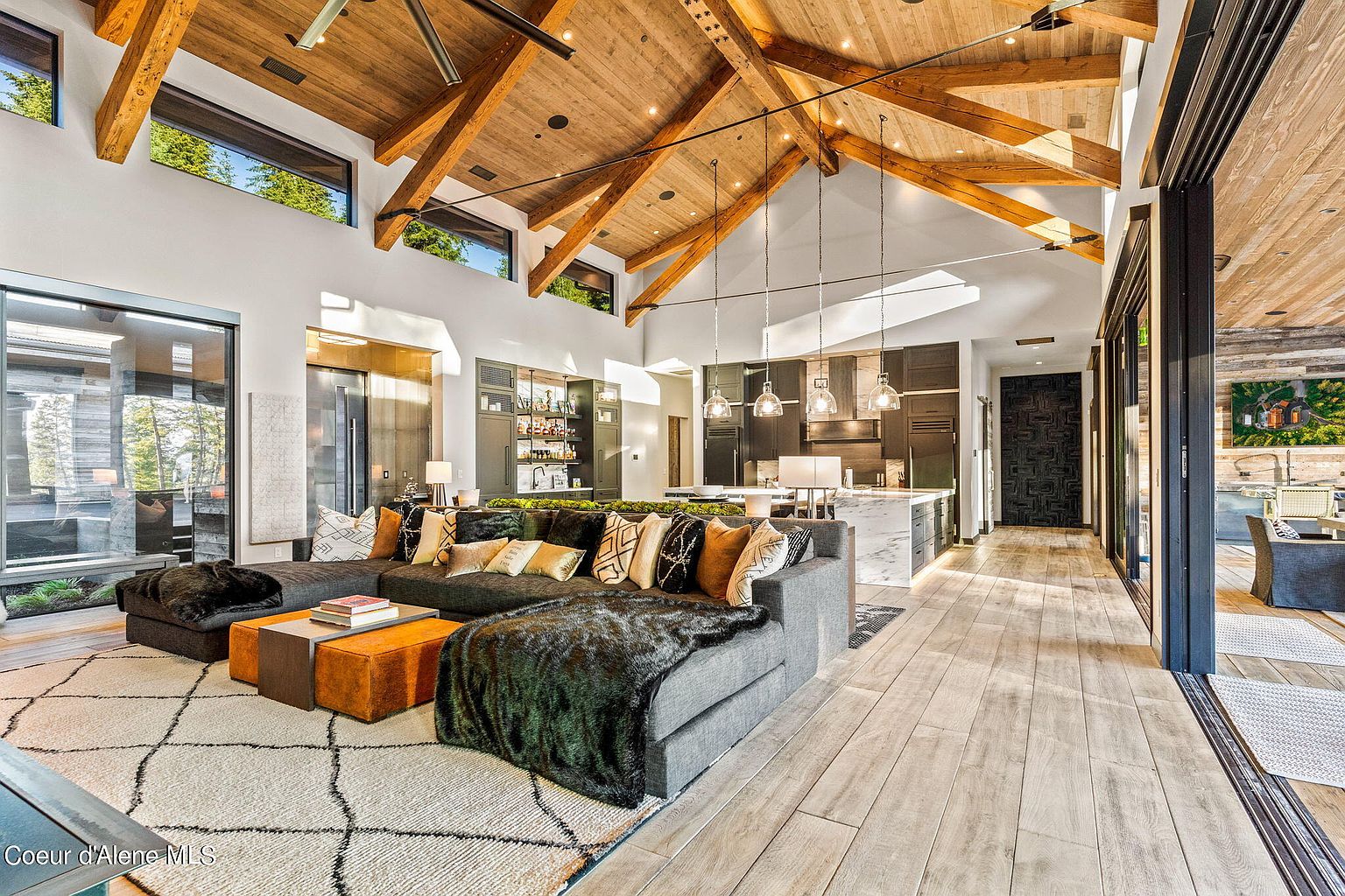 This is an interior shot of a luxurious living room that seamlessly blends into a modern kitchen. The living room features a large sectional sofa adorned with decorative pillows and throws, set upon a patterned rug. The kitchen boasts sleek cabinetry, a marble island, and pendant lighting, all under a high, vaulted ceiling with exposed wooden beams, creating an open and inviting space.
