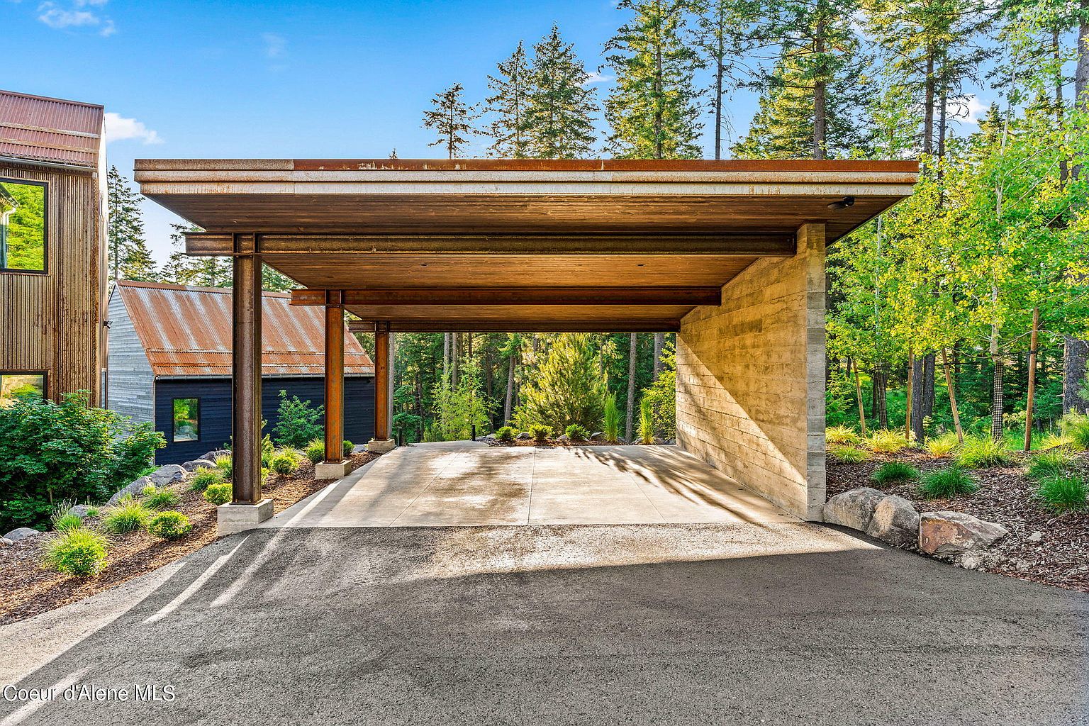 This image showcases a modern carport with a striking design, featuring a flat, cantilevered roof supported by steel beams and a concrete wall. The driveway leads into the carport, framed by lush greenery and mature trees, creating a sense of privacy and integration with nature. The architectural style is minimalist and contemporary, emphasizing clean lines and natural materials.