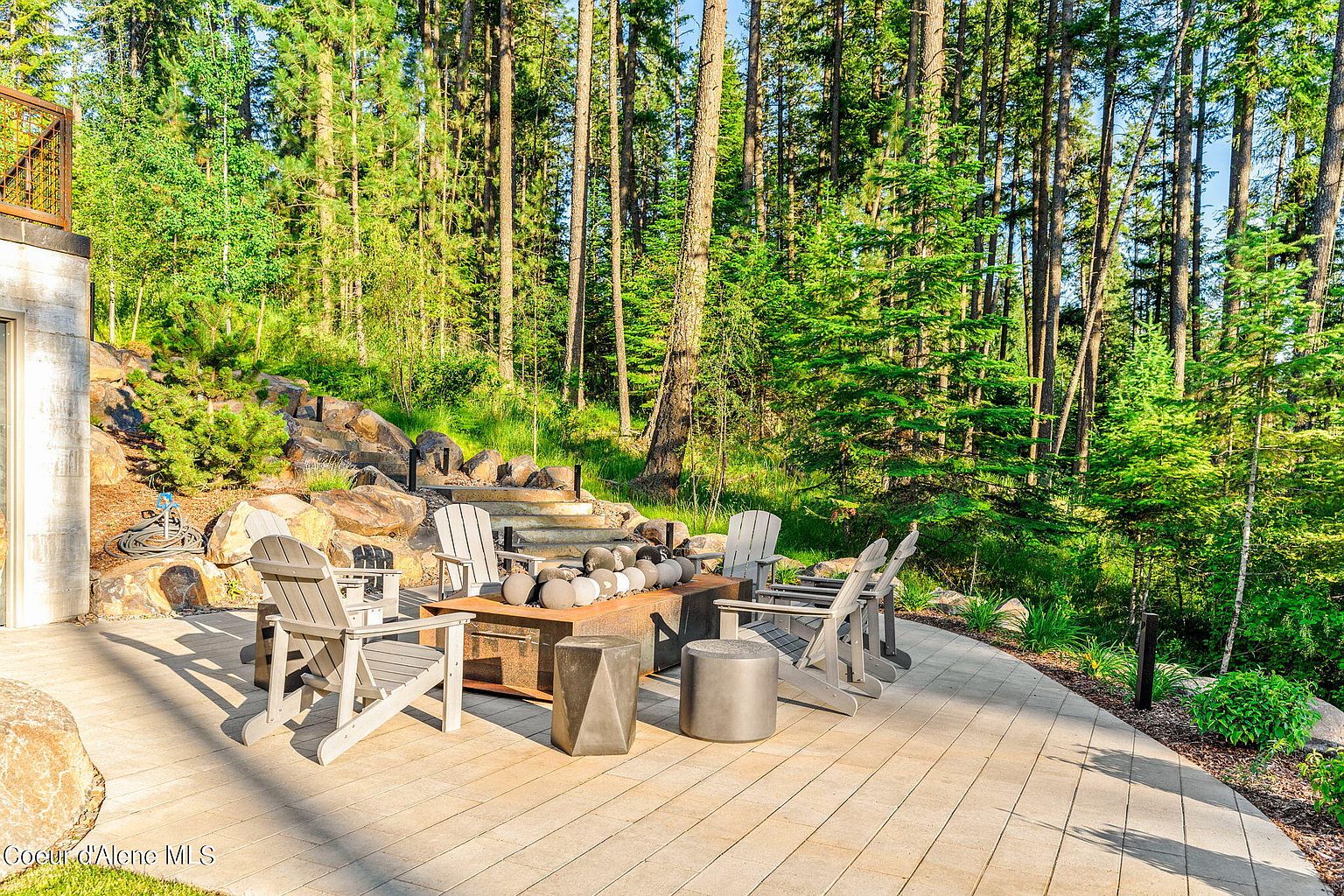 This image showcases a beautifully designed outdoor patio area, perfect for relaxation and entertainment. The patio features a modern fire pit surrounded by comfortable Adirondack chairs and stylish side tables, all set on a paved surface. The backdrop includes lush greenery and tall trees, creating a serene and private atmosphere, enhancing the property's appeal for outdoor living.