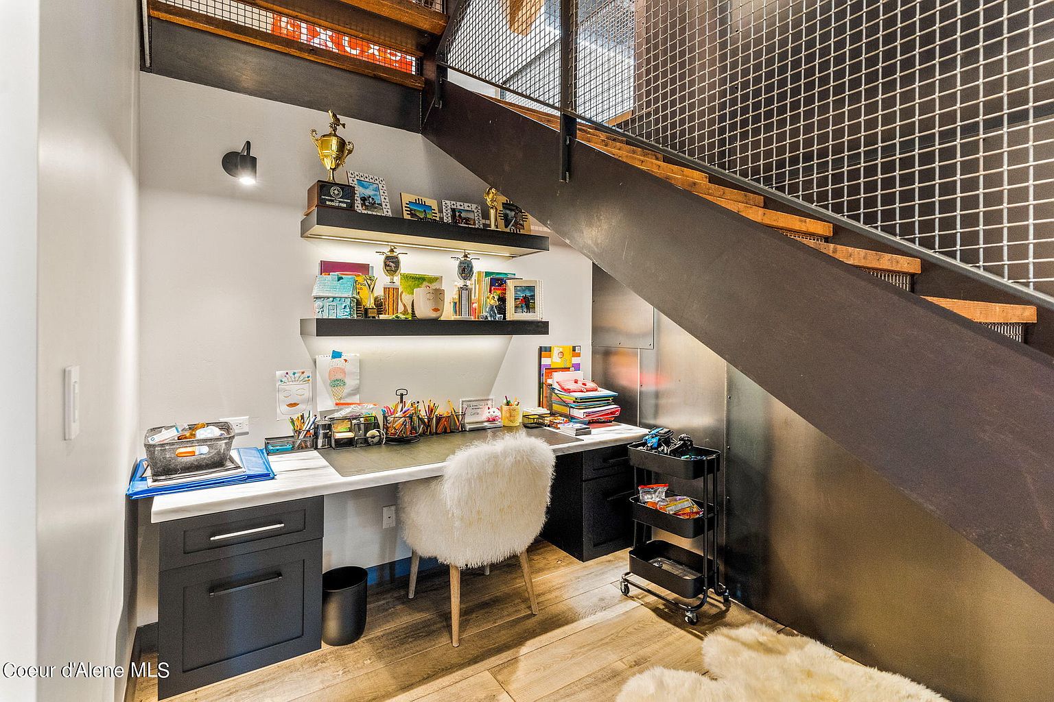 This interior shot showcases a cozy office space nestled under a staircase. The area features a desk with a white marble top and dark cabinetry, complemented by open shelving displaying trophies and decorative items. A fluffy white chair adds a touch of comfort, while a rolling cart provides additional storage, creating a functional and stylish workspace.