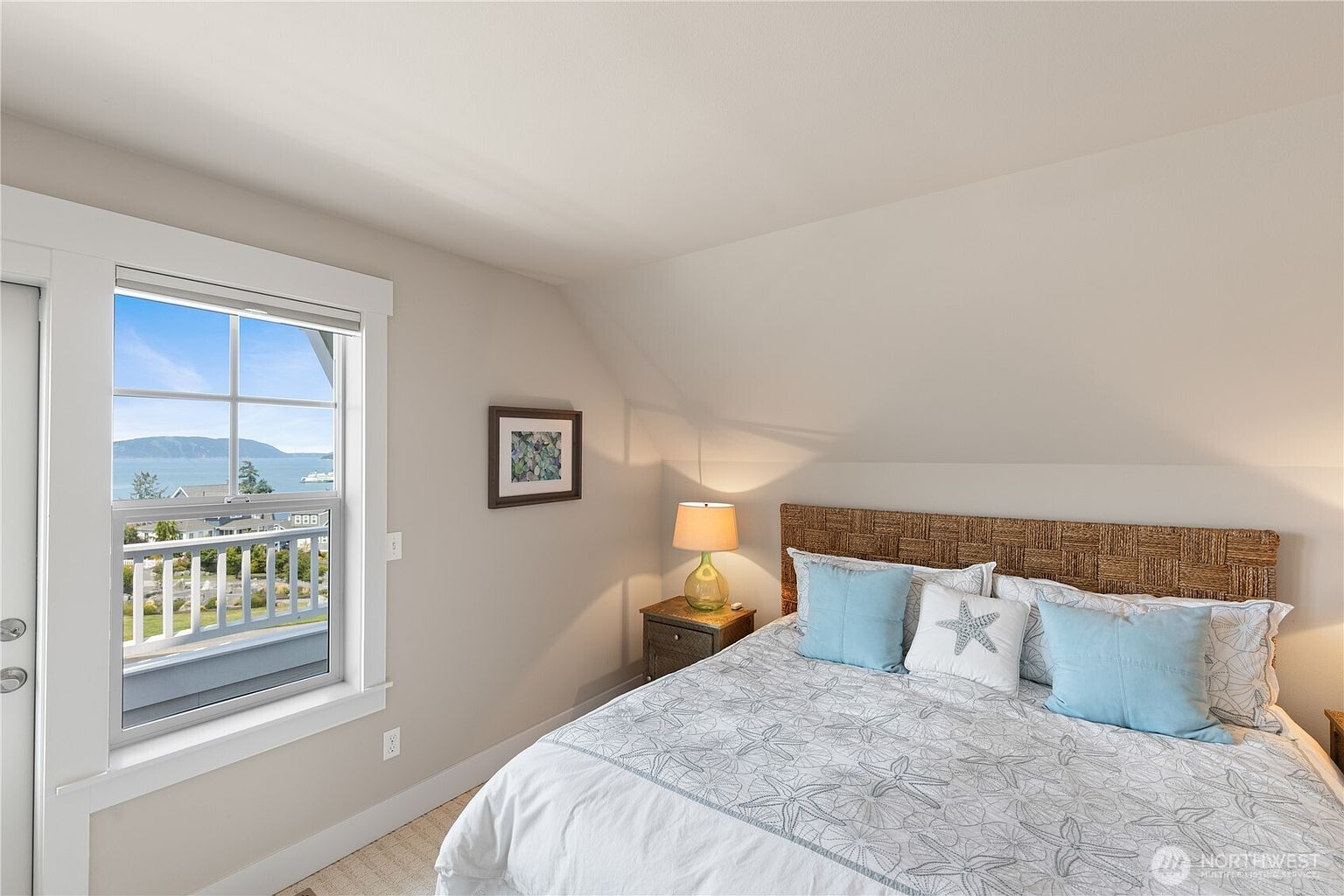 This cozy bedroom features a woven headboard and a bed dressed in a light-colored, starfish-patterned quilt with blue accent pillows. A window to the left provides a scenic view of the water and a balcony, while a bedside table with a lamp adds a warm, inviting touch. The room has a sloped ceiling, creating a comfortable and intimate atmosphere.