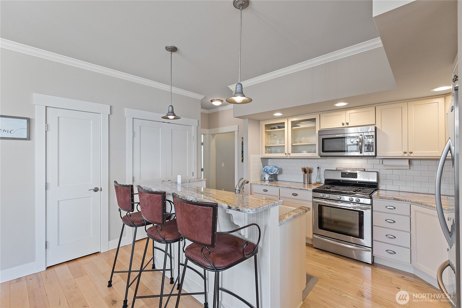 This bright kitchen features a functional layout with a breakfast bar island, light-toned cabinetry, and stainless steel appliances. The space is illuminated by pendant lighting over the counter and recessed ceiling lights, creating an inviting atmosphere. A neutral color palette and light wood flooring provide a clean, modern aesthetic suitable for daily living.