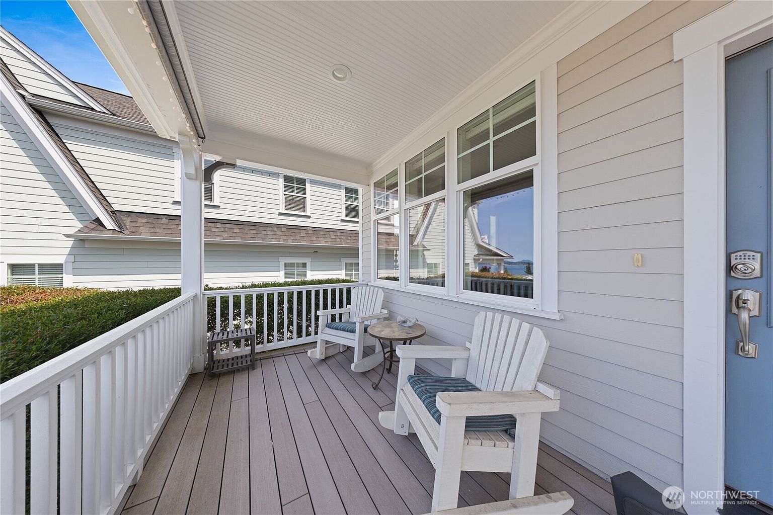 This inviting covered front porch features light-colored horizontal siding and a clean, coastal-inspired aesthetic. Two white wooden rocking chairs with striped cushions are positioned around a small round table, creating a cozy seating area. The space is framed by a white railing and large windows that reflect the surrounding neighborhood and a glimpse of the water.