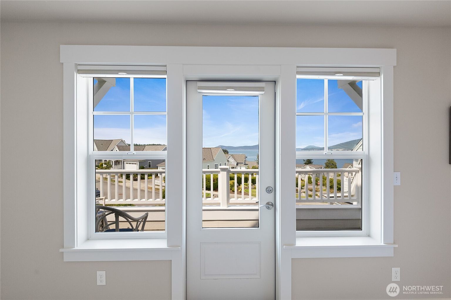This interior view features a central glass-paned door flanked by two large windows, creating a bright and airy focal point. The white-trimmed frames contrast against the neutral-toned walls, while the windows offer a clear view of an outdoor deck, neighboring homes, and the distant water. The perspective is straight-on, emphasizing the symmetry and the connection between the indoor living space and the scenic exterior.