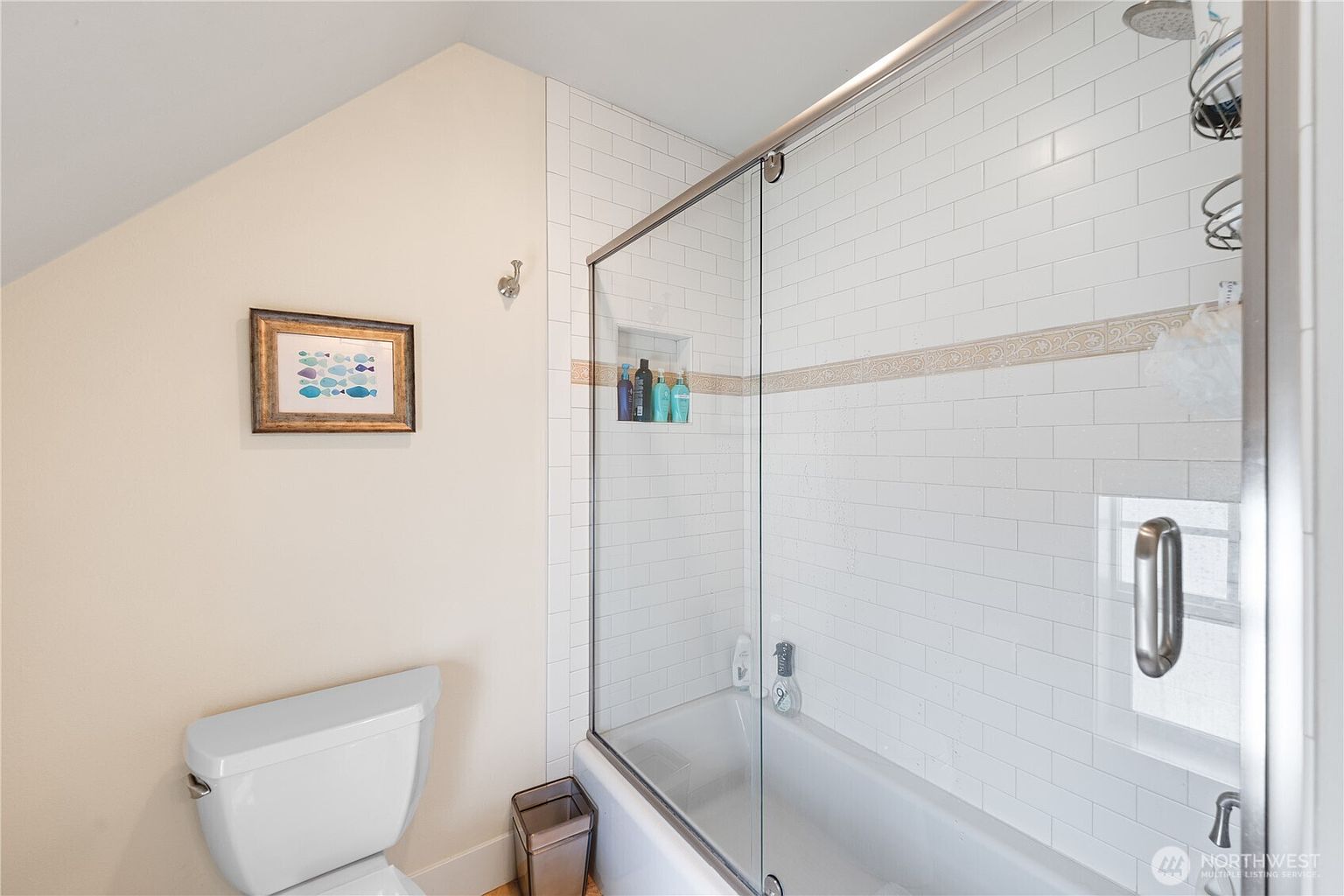 This bathroom features a shower-tub combination enclosed by a sliding glass door with a chrome frame. The shower walls are finished with white subway tiles and a decorative horizontal accent band, while a recessed niche provides convenient storage. A white toilet sits adjacent to the shower, and a framed fish-themed artwork hangs on the wall, adding a touch of character to the space.