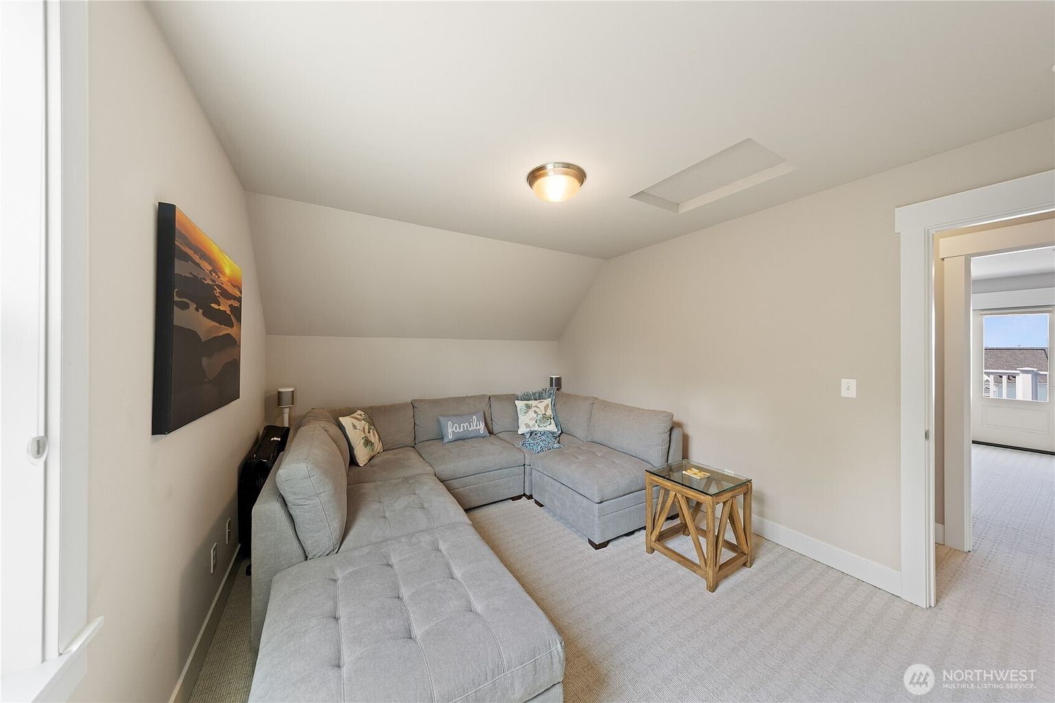 This cozy attic living space features a large, light gray sectional sofa tucked under a sloped ceiling, creating an intimate lounge area. A small glass-topped side table with a wooden base sits beside the sofa, while a piece of landscape art hangs on the wall. The room is carpeted in a neutral tone and includes a ceiling-mounted light fixture, with a doorway leading to an adjacent room visible on the right.