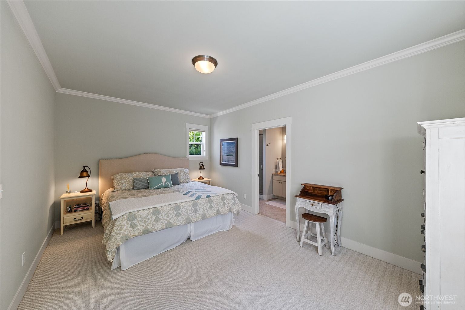 This primary bedroom features a neutral color palette with light-toned walls and plush carpeting, creating a serene and inviting atmosphere. A large bed with a beige upholstered headboard serves as the focal point, flanked by two matching nightstands with lamps. To the right, a small wooden writing desk with a stool sits near the doorway leading into an en-suite bathroom, while a white armoire provides additional storage.