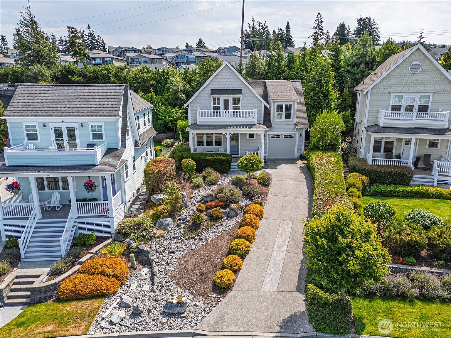 This elevated perspective captures a charming suburban street scene featuring three distinct, multi-story homes with classic architectural details like balconies and gabled roofs. The central property showcases a meticulously landscaped front yard with a dry creek bed design, stone accents, and a concrete driveway leading to an attached garage. The neighborhood is lush with mature trees and greenery, creating a serene and well-maintained residential atmosphere.