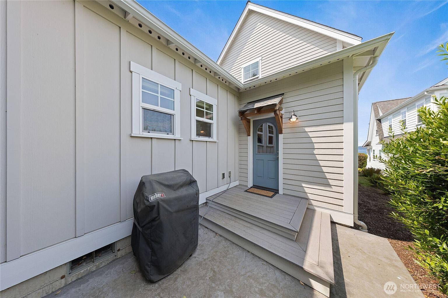 This exterior view features a side entryway with a light blue door sheltered by a small wooden-bracketed awning. A set of wooden steps leads up to the door, situated next to a covered grill on a concrete patio area. The home is finished with a mix of vertical board-and-batten and horizontal lap siding, creating a charming, cottage-like aesthetic.