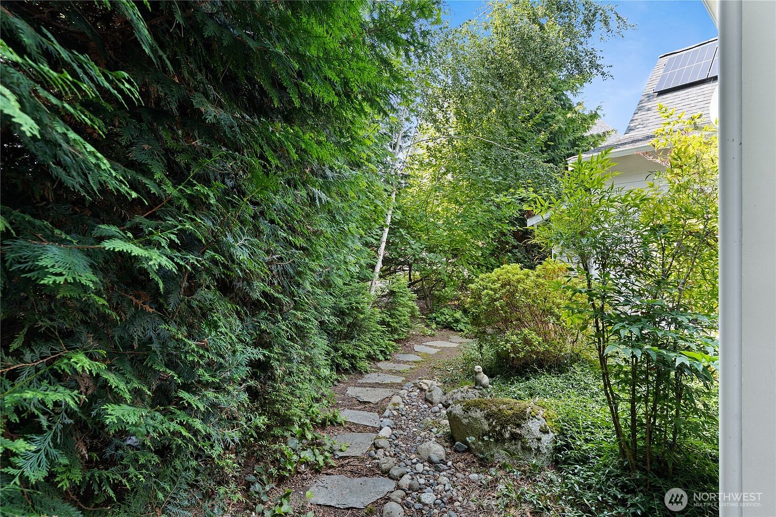 This image captures a serene, shaded garden path winding alongside the side of a white-sided home. The walkway is composed of irregular stone pavers set into the earth, bordered by a lush, dense hedge of evergreen trees on the left and a variety of shrubs and a mossy boulder on the right. The perspective is from the edge of the path, looking toward the rear of the property where sunlight filters through the mature trees.