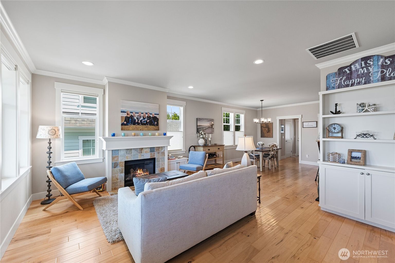 This open-concept living area features light wood flooring and a neutral color palette, creating a bright and inviting atmosphere. A central fireplace with a decorative tile surround serves as a focal point, flanked by two blue accent chairs and a comfortable sofa. The space flows seamlessly into a dining area in the background, with built-in shelving providing organized storage and display space.