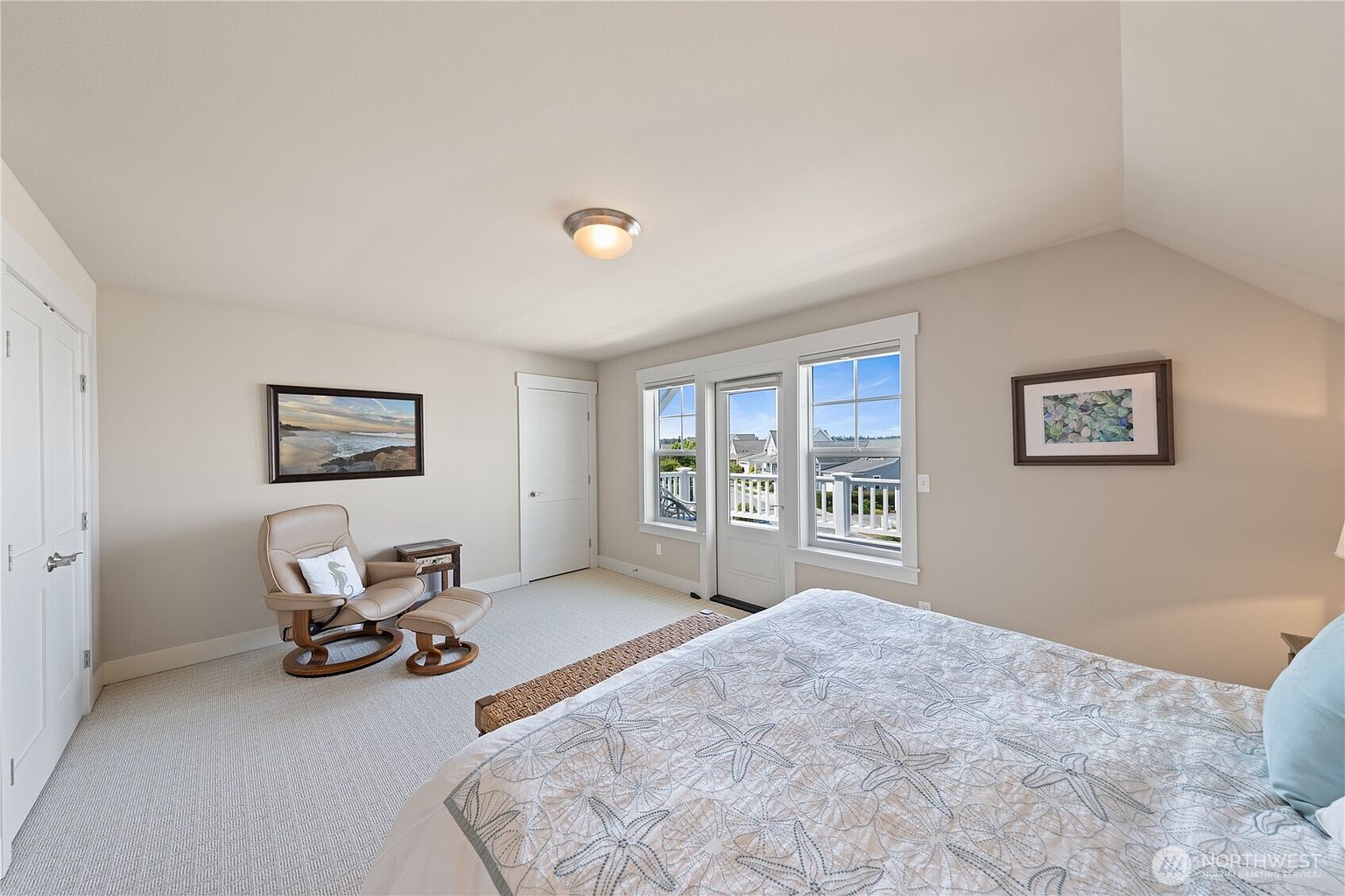 This bright and airy primary bedroom features neutral walls, light-colored carpeting, and a bed adorned with a coastal-themed starfish patterned quilt. A comfortable recliner with an ottoman sits in the corner near a framed beach landscape, while a glass-paned door provides access to an outdoor balcony. The room is illuminated by a central ceiling light fixture and natural light streaming through the windows.