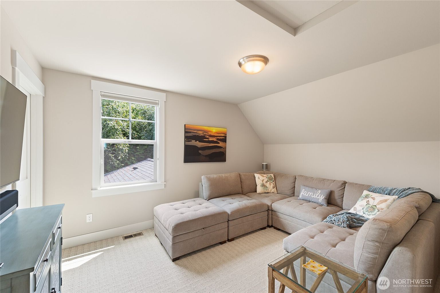 This cozy living room features a large, neutral-toned sectional sofa with an attached ottoman, creating a comfortable seating area under a sloped ceiling. A window provides natural light and a view of the outdoors, while a piece of landscape art hangs on the wall above the sofa. The room is finished with light-colored carpeting and a small glass-topped side table, offering a relaxed and inviting atmosphere.
