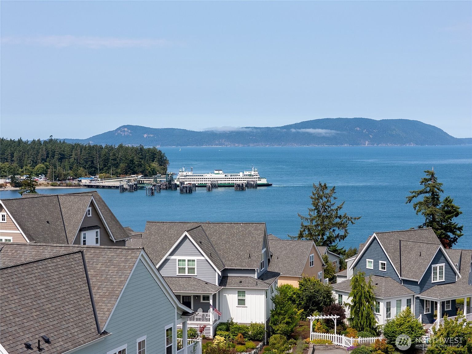 This elevated perspective captures a scenic coastal neighborhood featuring several charming, multi-story homes with shingled roofs and varied siding colors. In the background, a large ferry is docked at a pier, set against a backdrop of deep blue water and distant, forested mountains under a clear sky. The composition emphasizes the desirable waterfront location and the peaceful, residential atmosphere of the community.