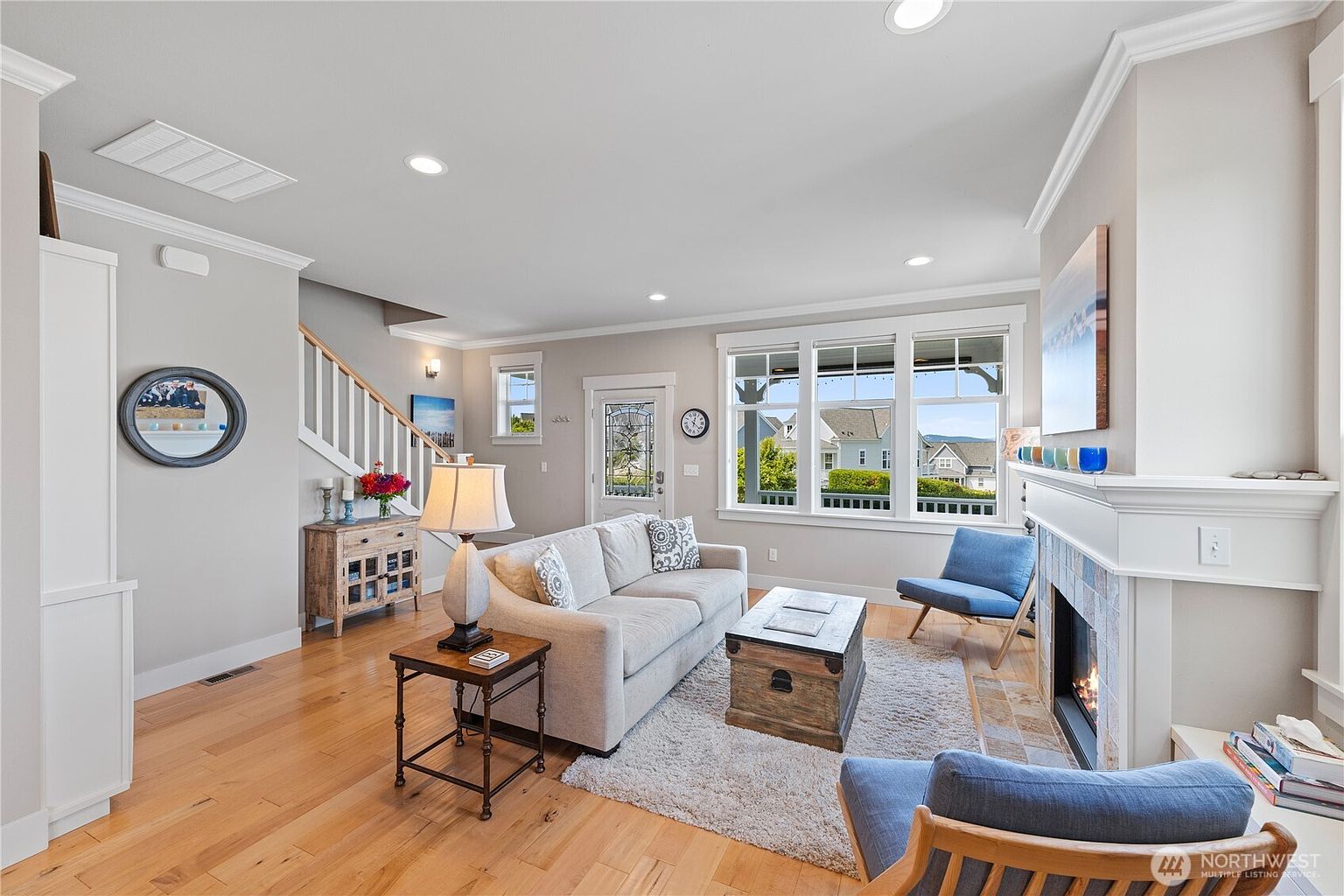 This bright and inviting living room features light wood flooring, a neutral-toned sofa, and a central wooden trunk coffee table resting on a textured area rug. A fireplace with a white mantel and tiled surround serves as a focal point, complemented by two blue accent chairs and a side table with a lamp. The space is illuminated by recessed lighting and natural light streaming through a large window arrangement that offers a view of the neighborhood.