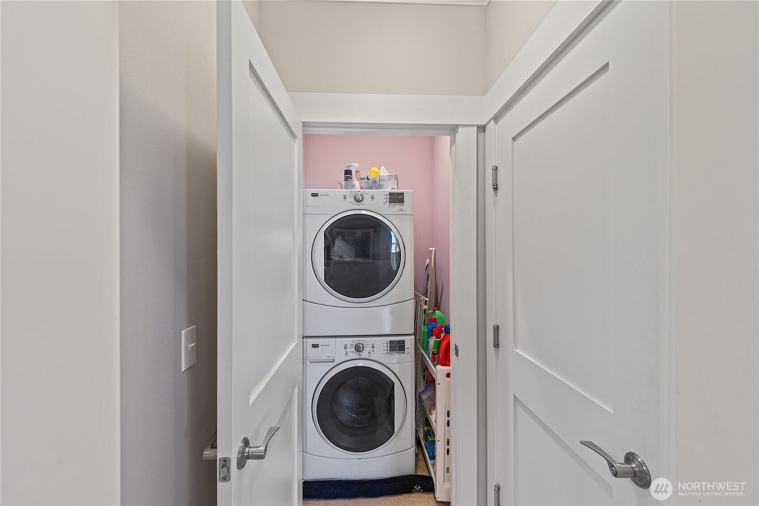 This compact laundry closet features a stacked white washer and dryer unit set against a soft pink wall. To the right of the appliances, a slim, multi-tiered rolling cart provides organized storage for cleaning supplies and laundry essentials. The space is accessed through double white doors, presenting a clean and functional utility area.