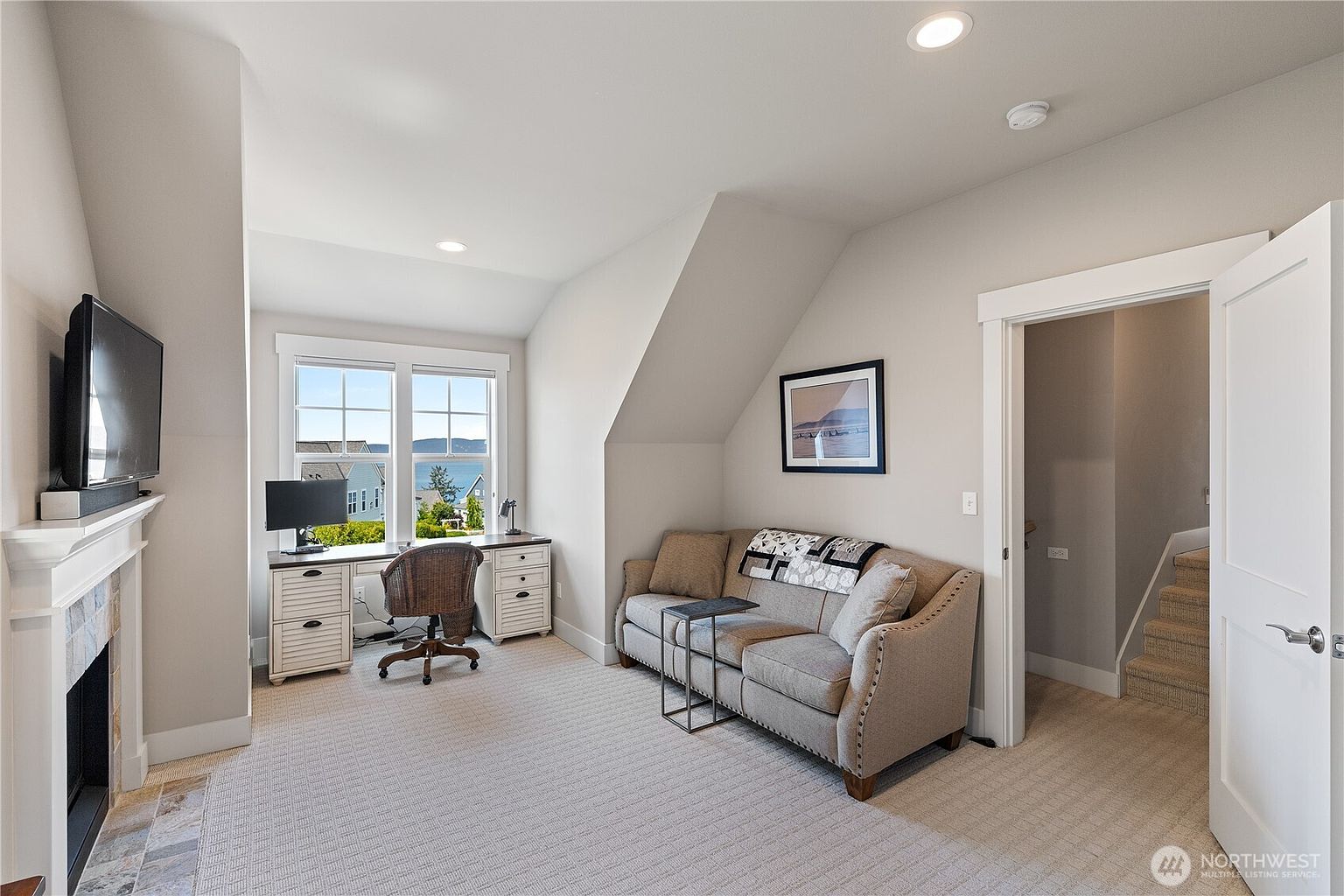 This versatile room functions as a home office and cozy lounge, featuring a desk positioned by a window with a scenic water view. A neutral-toned sofa with a patterned throw blanket sits opposite a fireplace with a white mantel, creating a comfortable and inviting atmosphere. The space is finished with light-colored carpeting and neutral wall tones, with a doorway leading to a carpeted staircase.