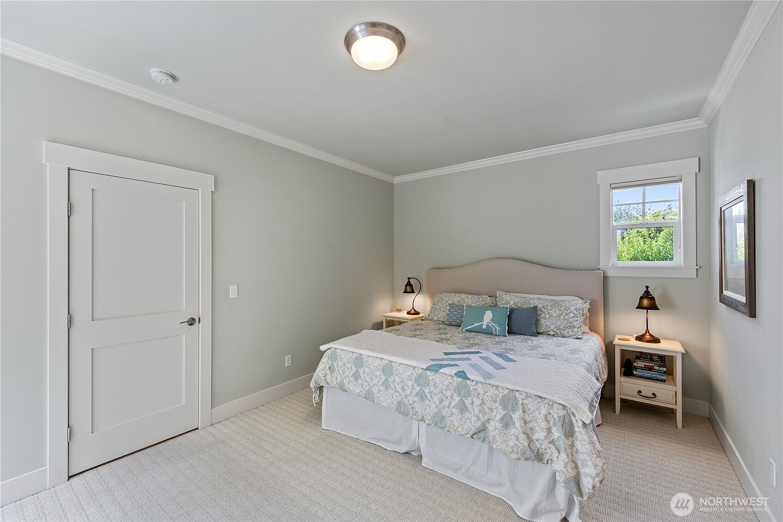 This bright and neutral bedroom features a queen-sized bed with a light-colored upholstered headboard and patterned bedding. The room is illuminated by a central ceiling light fixture and two matching bedside lamps, with a single window providing natural light. The space is finished with light-toned walls, white trim, and wall-to-wall carpeting, creating a clean and inviting atmosphere.