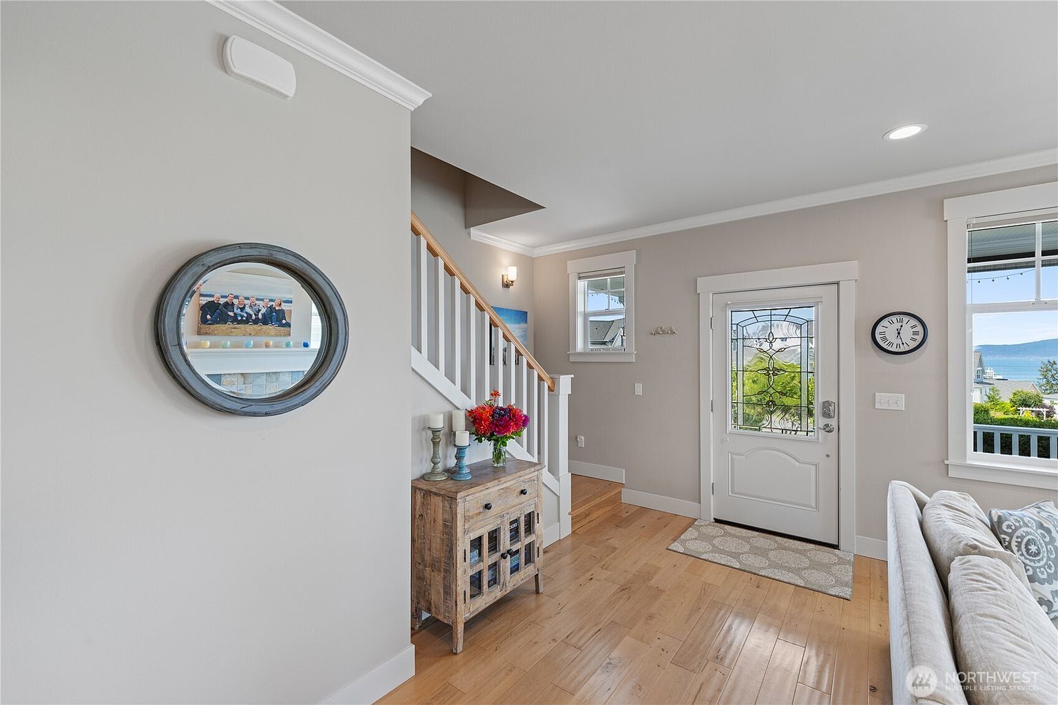 This bright entryway features light wood flooring and neutral walls, creating an inviting and airy atmosphere. A wooden console table sits beneath a staircase with white railings, while a round mirror on the wall reflects the living space. The front door, accented with decorative glass, allows natural light to flow into the home, complemented by a view of the outdoors through a nearby window.