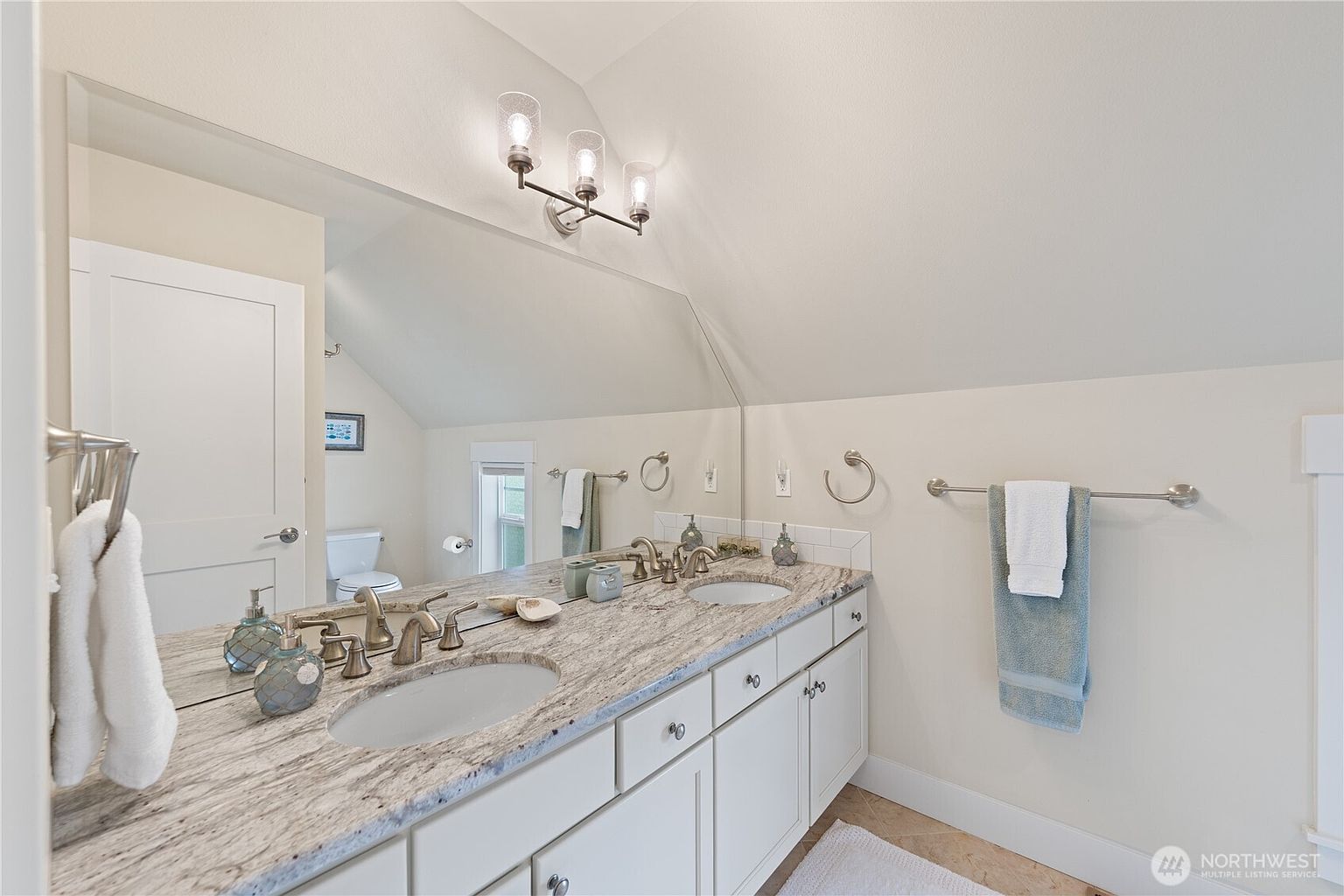 This bright bathroom features a double-sink vanity with a light-colored stone countertop and white cabinetry. A large, angled mirror reflects the space, including a glimpse of the toilet area and a window, while a sloped ceiling adds architectural interest. The room is finished with neutral wall tones and polished metal fixtures, creating a clean and functional aesthetic.