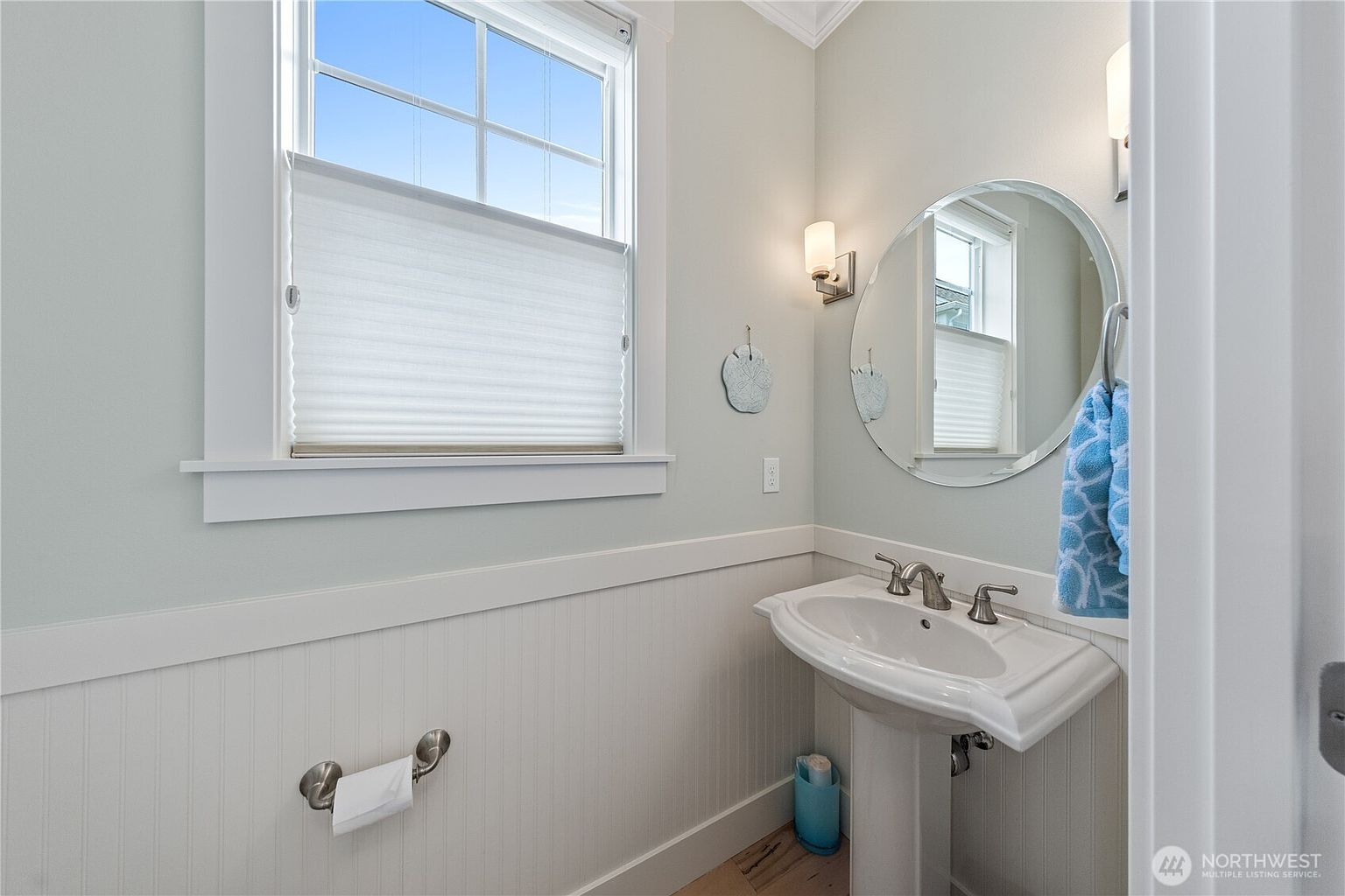 This bright and airy powder room features a classic pedestal sink and a round mirror mounted on a light-colored wall. The space is accented with white wainscoting, a window with a pleated shade, and two wall-mounted light fixtures. The overall aesthetic is clean, traditional, and welcoming.