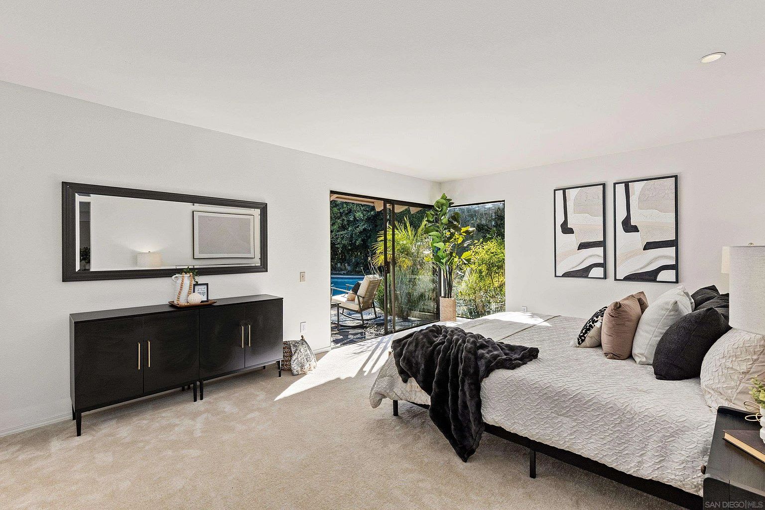 This is a bright and airy primary bedroom featuring a modern aesthetic. The room includes a large bed with multiple pillows and a throw blanket, a dark wood dresser with a mirror above it, and sliding glass doors that lead to an outdoor area with a pool. The walls are painted a clean white, and the room is well-lit with natural light.