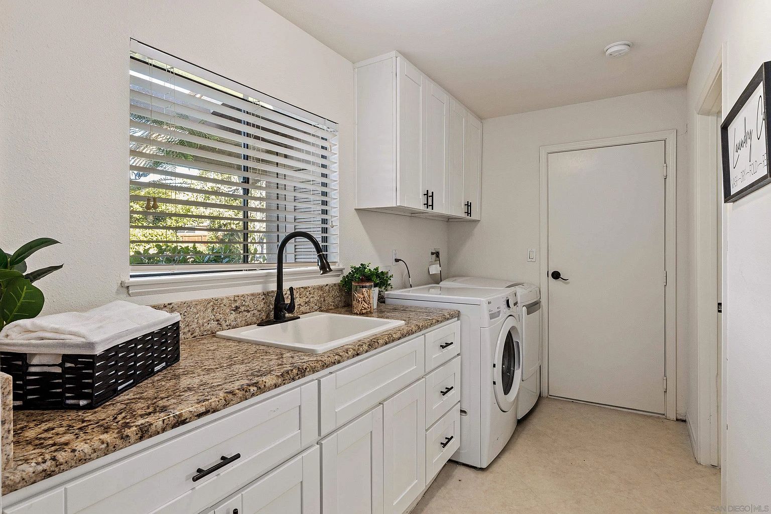 This is a well-lit laundry room featuring white cabinetry with black hardware, granite countertops, and a stainless steel sink with a black faucet. A front-loading washer and dryer are present, and a window with blinds provides natural light. The room appears clean and functional, ideal for a modern home.