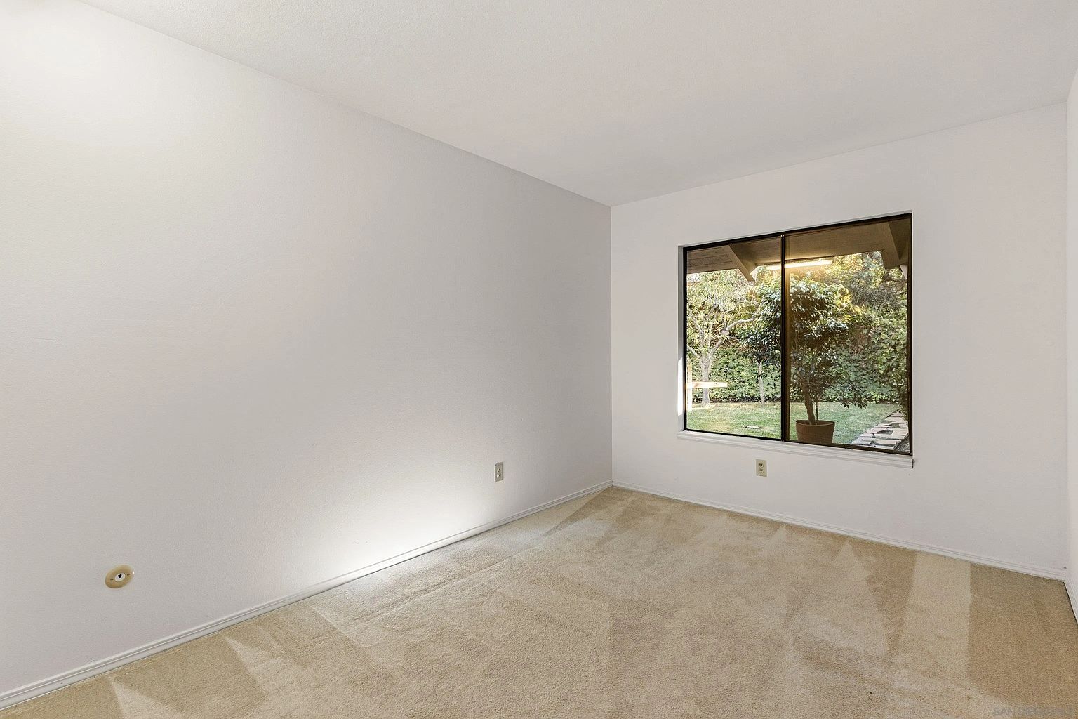 The image showcases a simple, carpeted bedroom with neutral tones. A window provides natural light and a view of the outdoors, featuring greenery and a glimpse of a garden. The room is sparsely furnished, emphasizing its potential for customization and highlighting the clean, minimalist aesthetic.