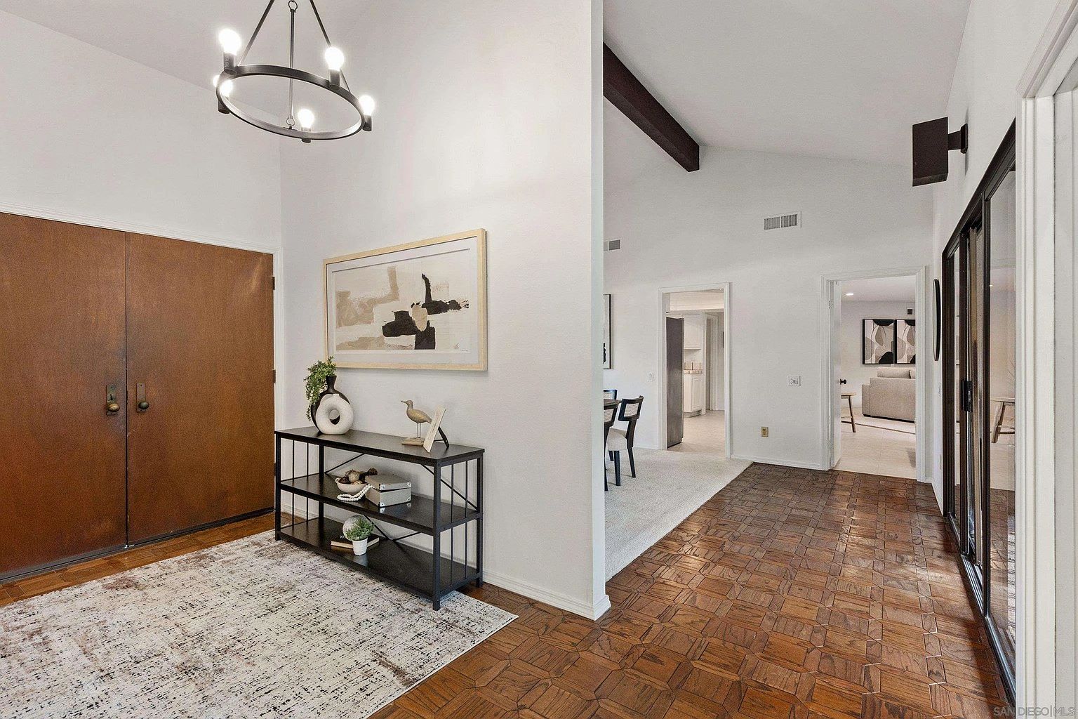 This interior shot showcases a well-lit hallway with a blend of modern and mid-century design elements. The entryway features a dark wood double door, a black metal console table with decorative items, and a framed abstract artwork. The flooring transitions from a patterned area rug to a parquet wood design, leading to other rooms visible through open doorways.