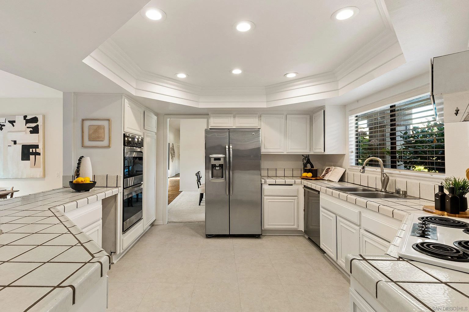 This is a well-lit kitchen with white cabinets, tiled countertops, and stainless steel appliances, including a double-door refrigerator and built-in ovens. The kitchen features a tiled floor and a window above the sink, providing natural light. The overall impression is a clean and functional space.