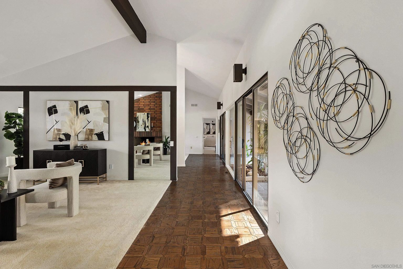 This interior shot showcases a well-lit hallway with a blend of carpet and wood flooring, leading to various rooms. The hallway features modern wall art, large glass windows offering natural light, and dark wood accents on the ceiling beams and door frames. The design creates a sense of spaciousness and contemporary elegance.