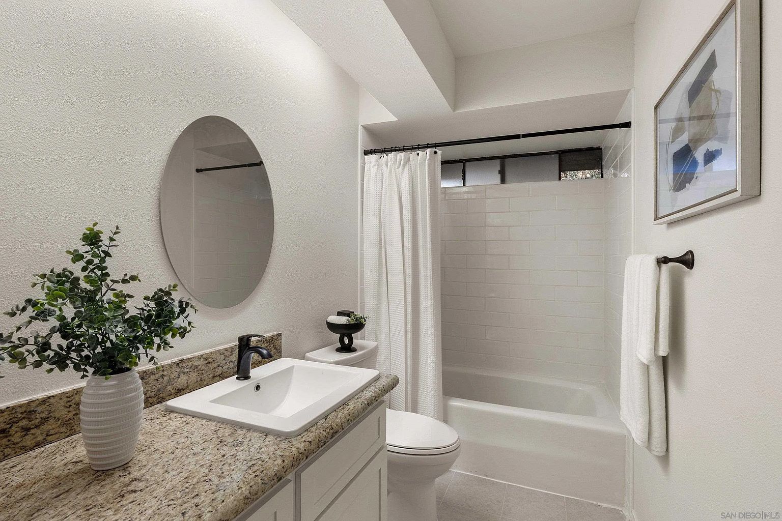 This is a well-lit bathroom featuring a granite countertop vanity with a white sink and a round mirror. A toilet sits next to the vanity, and a bathtub with a white shower curtain is visible in the background. The walls are painted white, and a framed piece of art hangs on the wall, creating a clean and modern aesthetic.